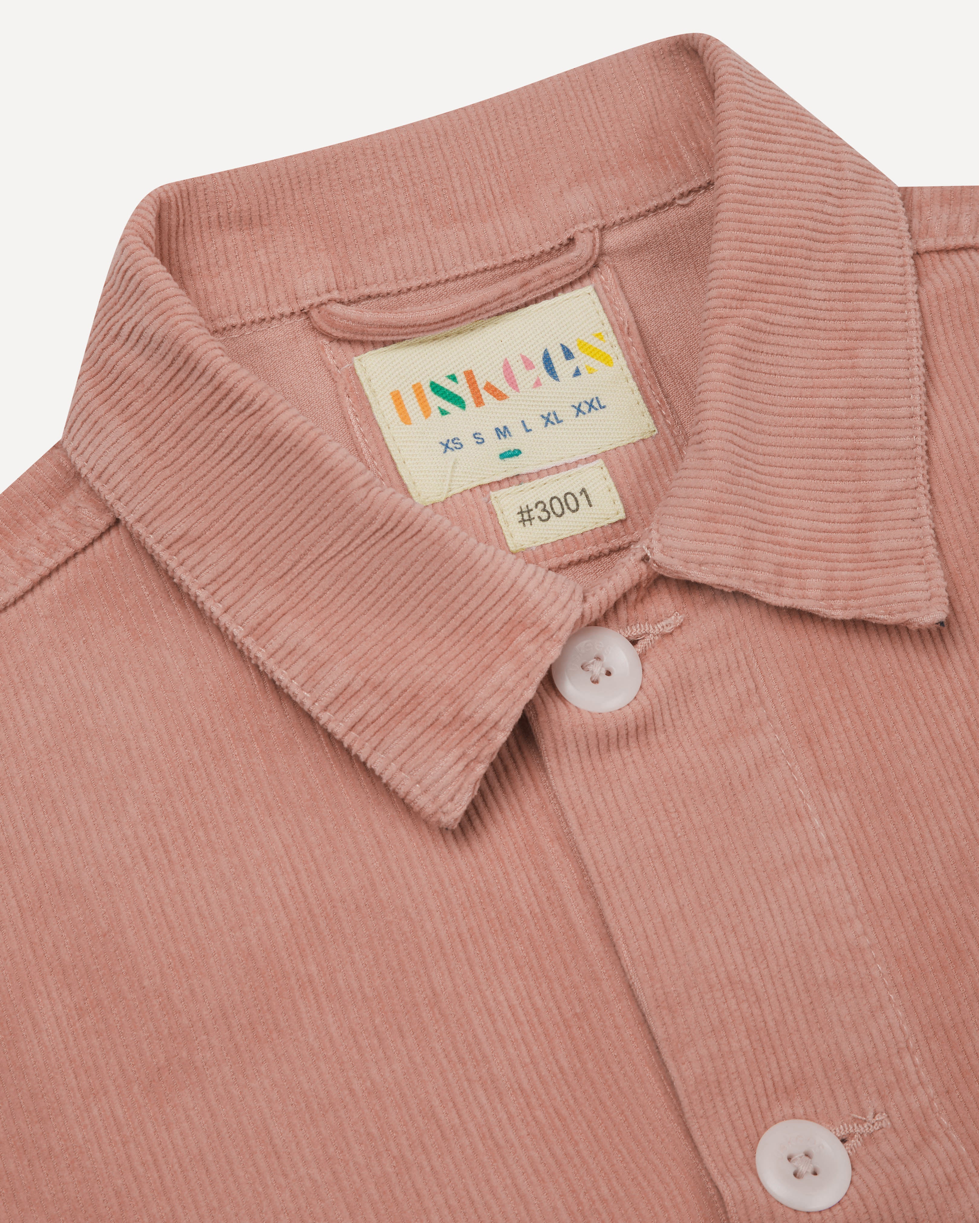 Front close up shot of pink, buttoned corduroy overshirt from Uskees. Clear view of collar, brand/size label and corozo buttons, 
