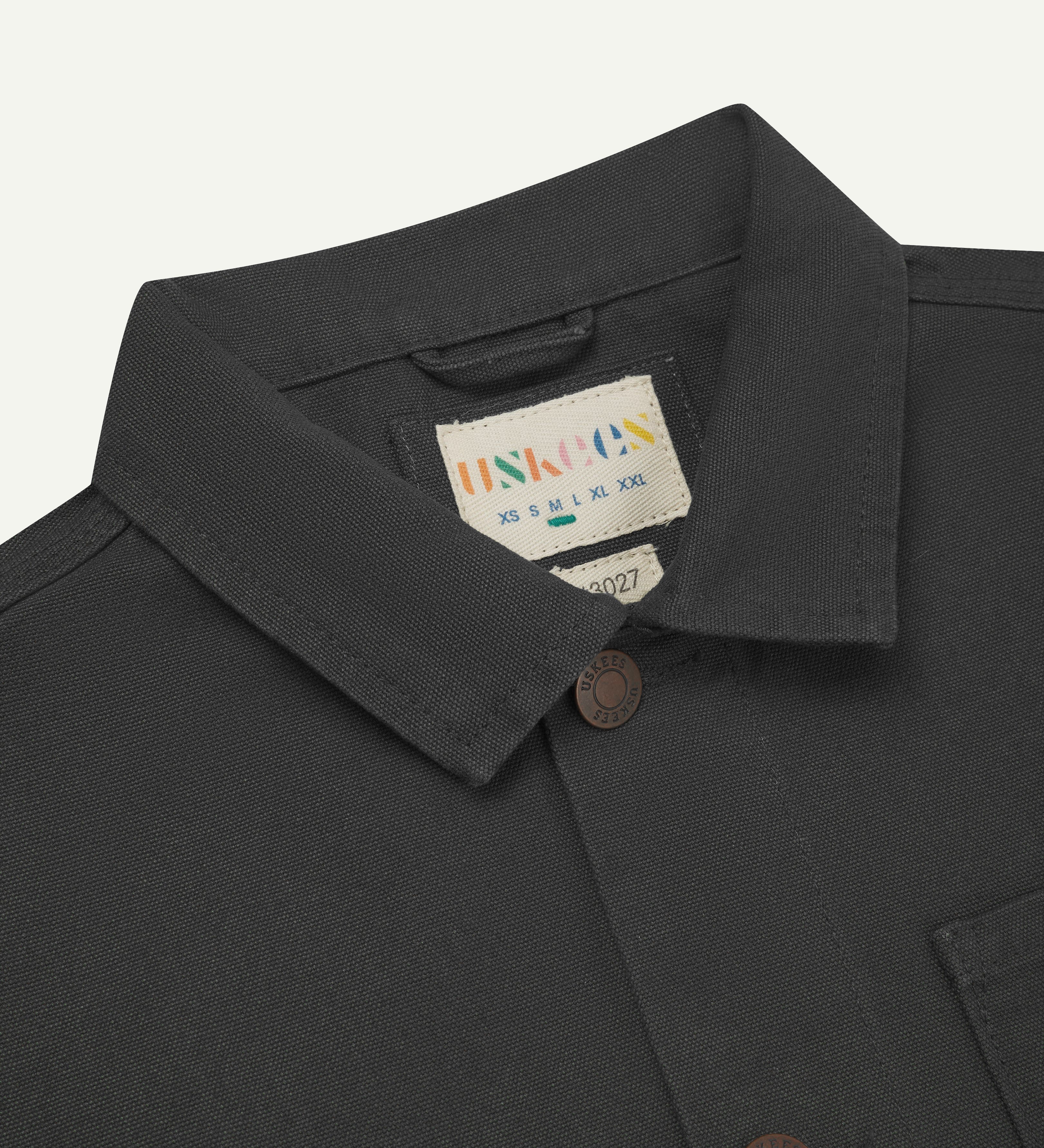 Front close-up view of uskees dark grey canvas men's overshirt presented buttoned up showing the metal buttons, collar and brand label.