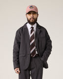 Front view of a bearded male model wearing the Uskees charcoal docklands blazer, matching trousers, a white shirt and a striped tie.