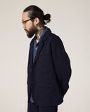 Side view of a bearded, bespectacled male model wearing the Uskees midnight blue docklands blazer, matching trousers and a blue shirt.