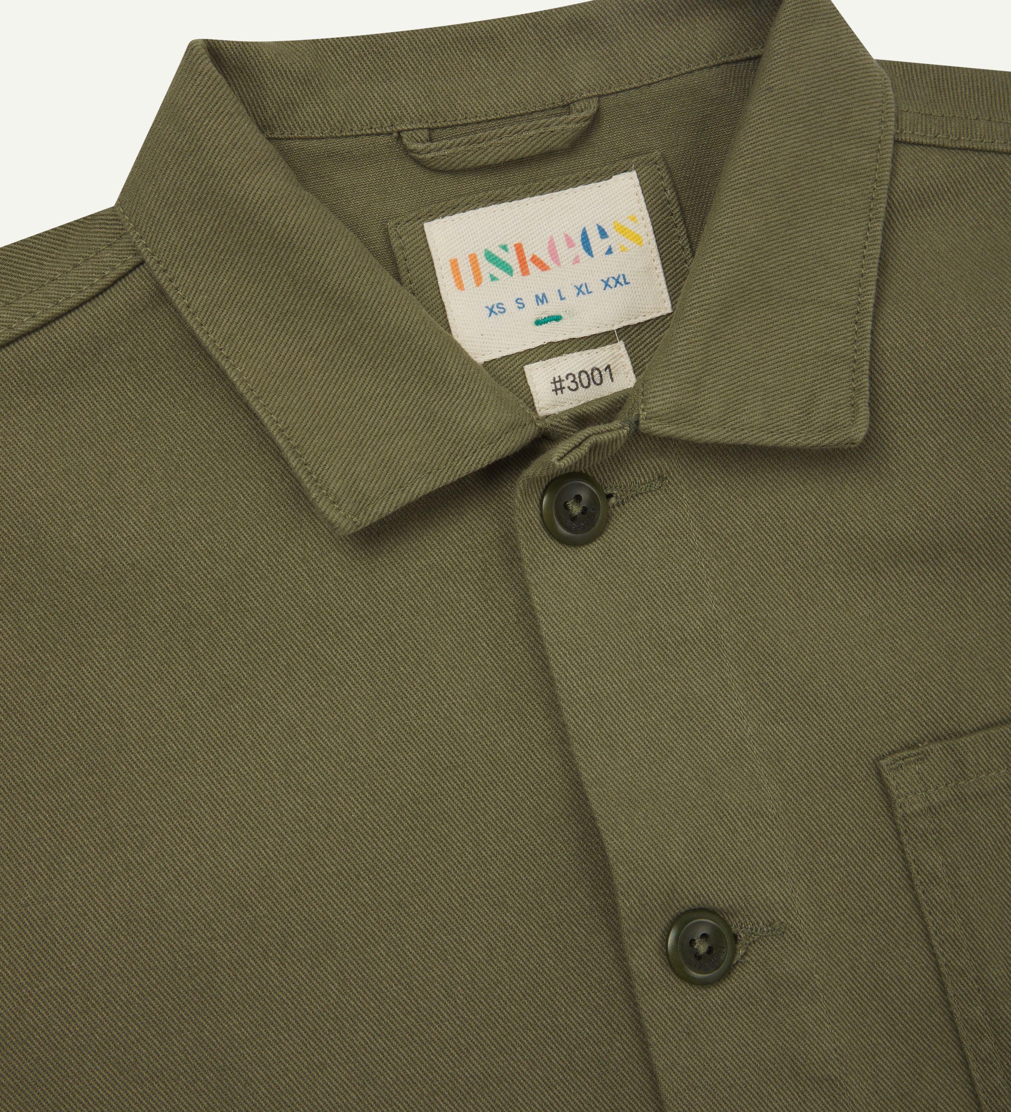 Close up of moss-green buttoned organic cotton-drill overshirt from Uskees. Clear view of collar and Uskees branding label.