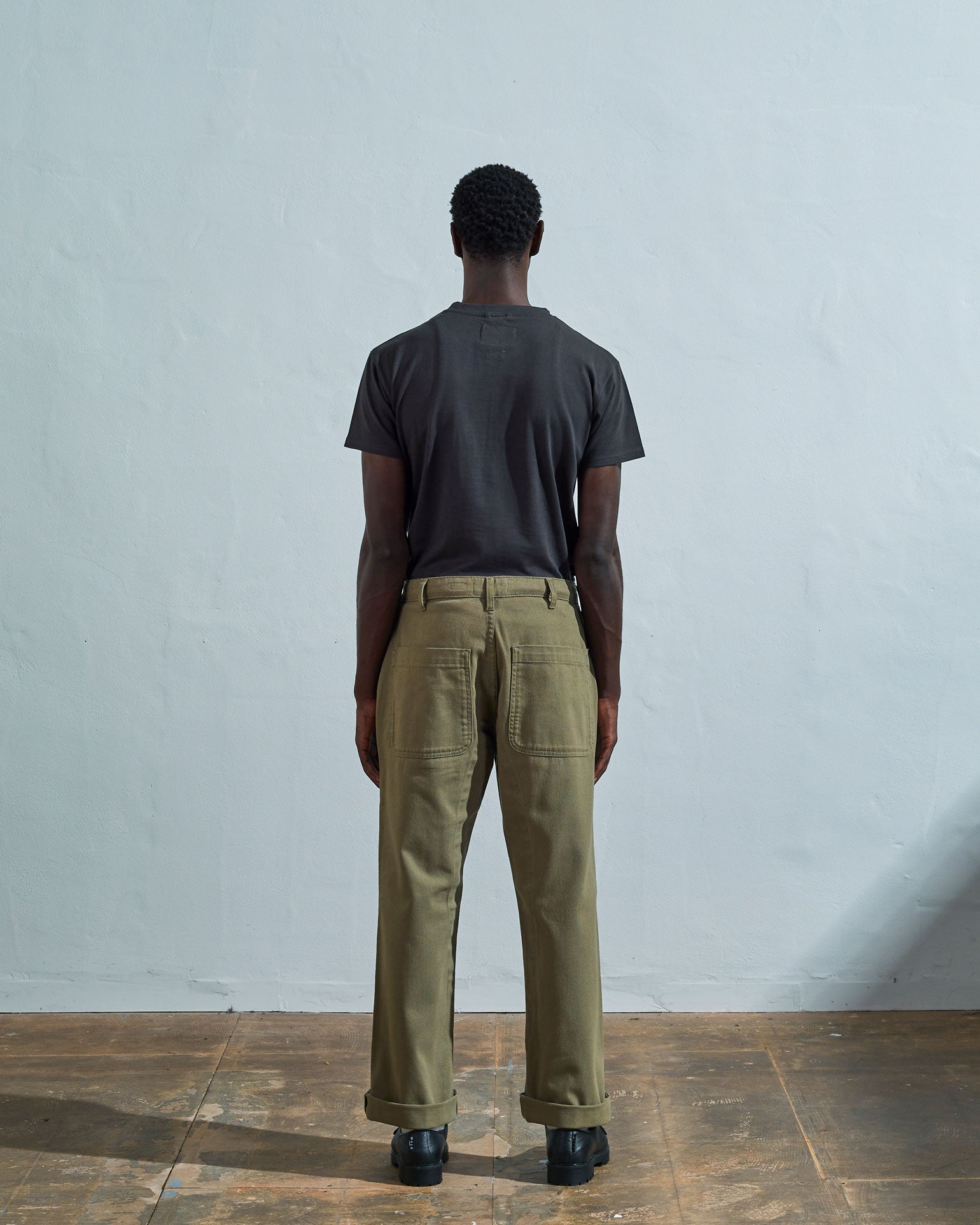 Full-length reverse view of model wearing #5016 drill commuter pants in moss, with focus on belt loops and large rear pockets.