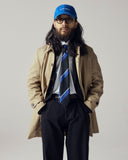 Male model with a beard wearing a blue Uskees baseball cap, glasses, beige car coat, midnight blue Uskees pants, and striped tie against a plain background