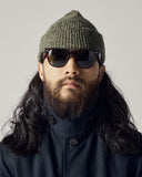 Head and shoulders shot of a male model wearing an Uskees midnight blue car coat, green knit beanie and sunglasses with a neutral background