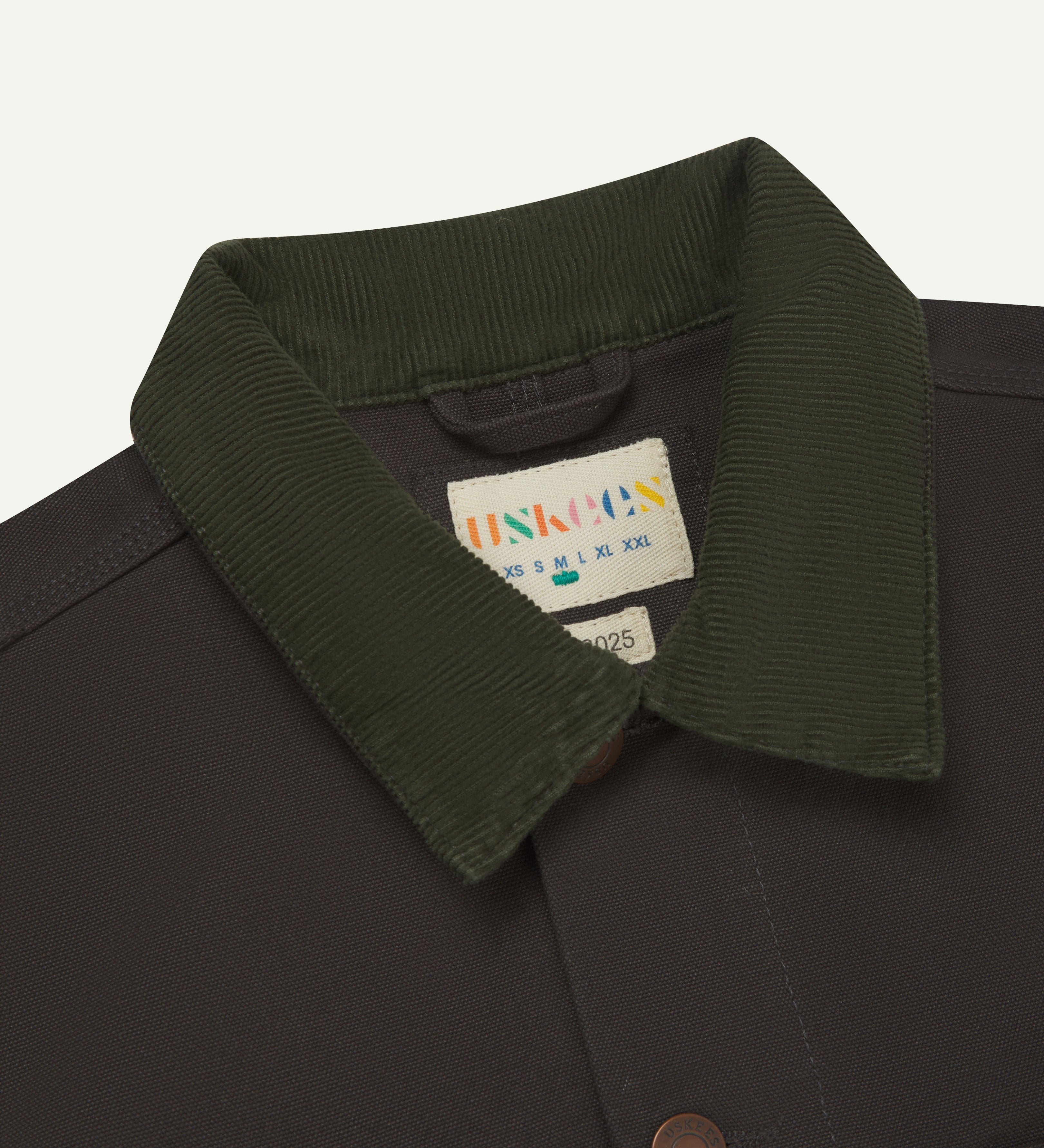 Front close-up shot of uskees cotton canvas chore jacket for men in dark grey. Clear view of the green corduroy collar, brand label and buttons