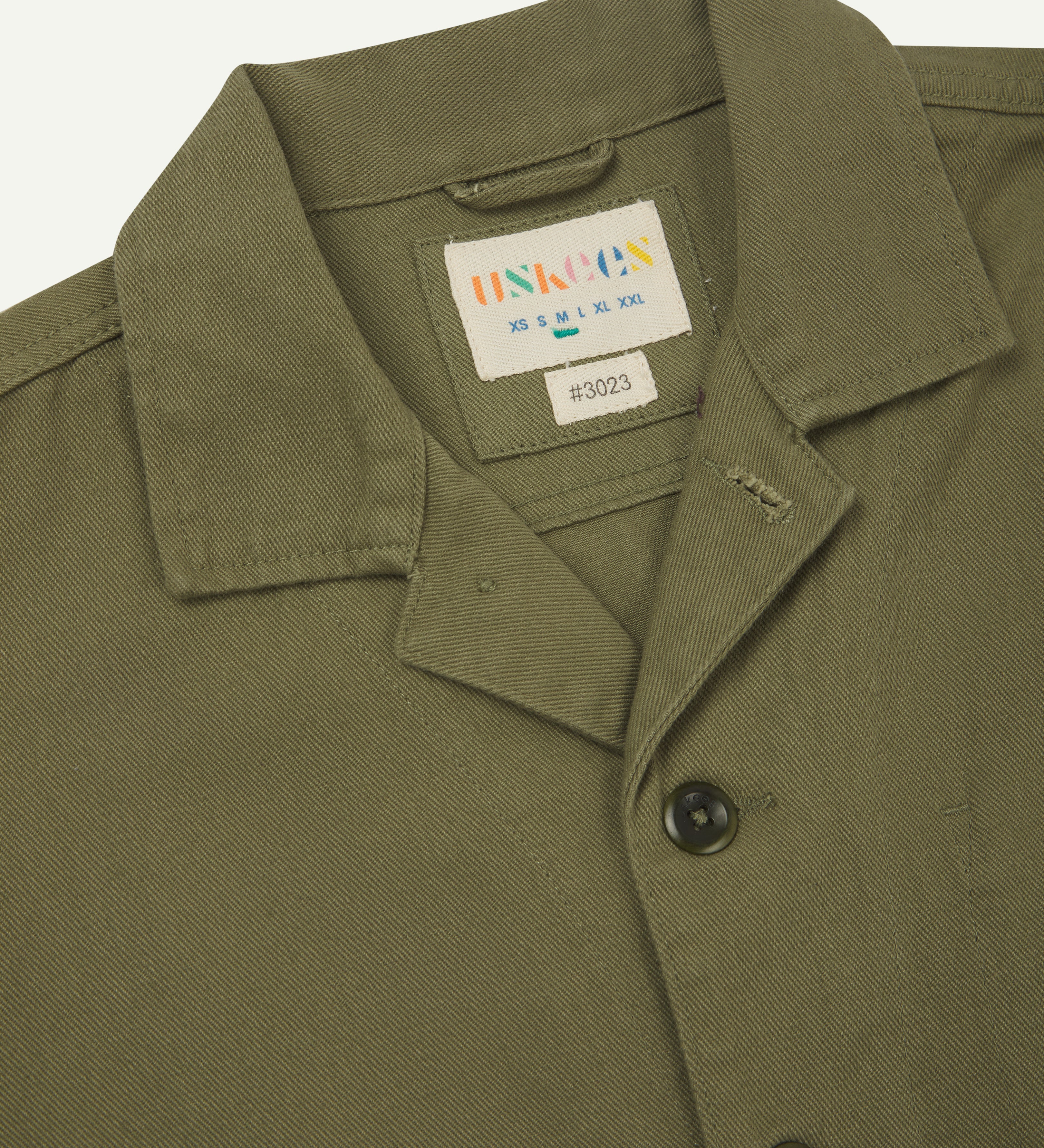 Front close view of moss green organic cotton drill commuter blazer with patch pockets showing collar, corozo buttons and Uskees branding label.