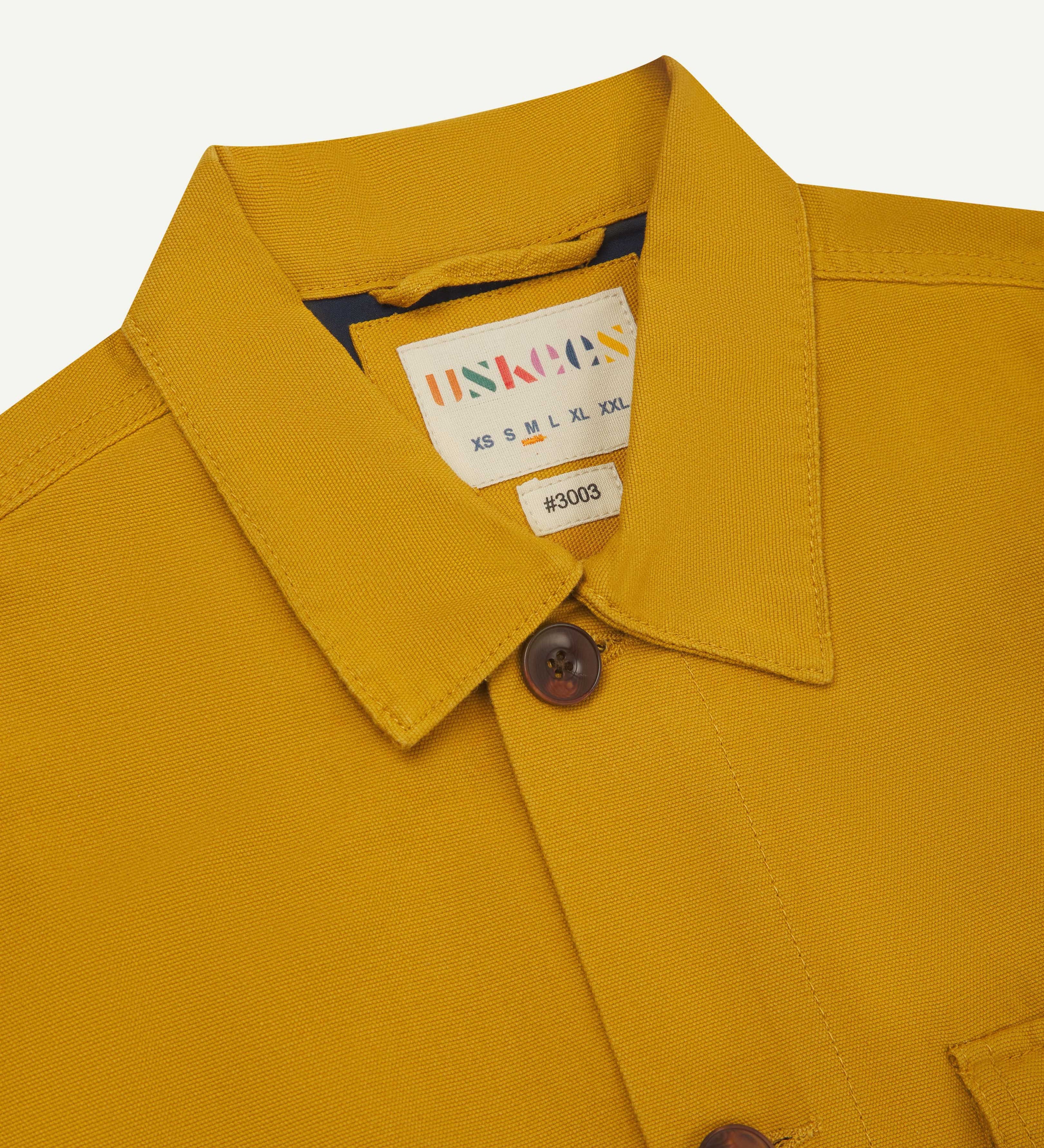 front detail view of uskees bright yellow workshirt for men showing brand label and blue yoke lining