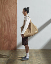 Full-length, front-view of model carrying Uskees #4001, large khaki tote bag over left shoulder. Paired with #3002, light grey zip-front jacket.