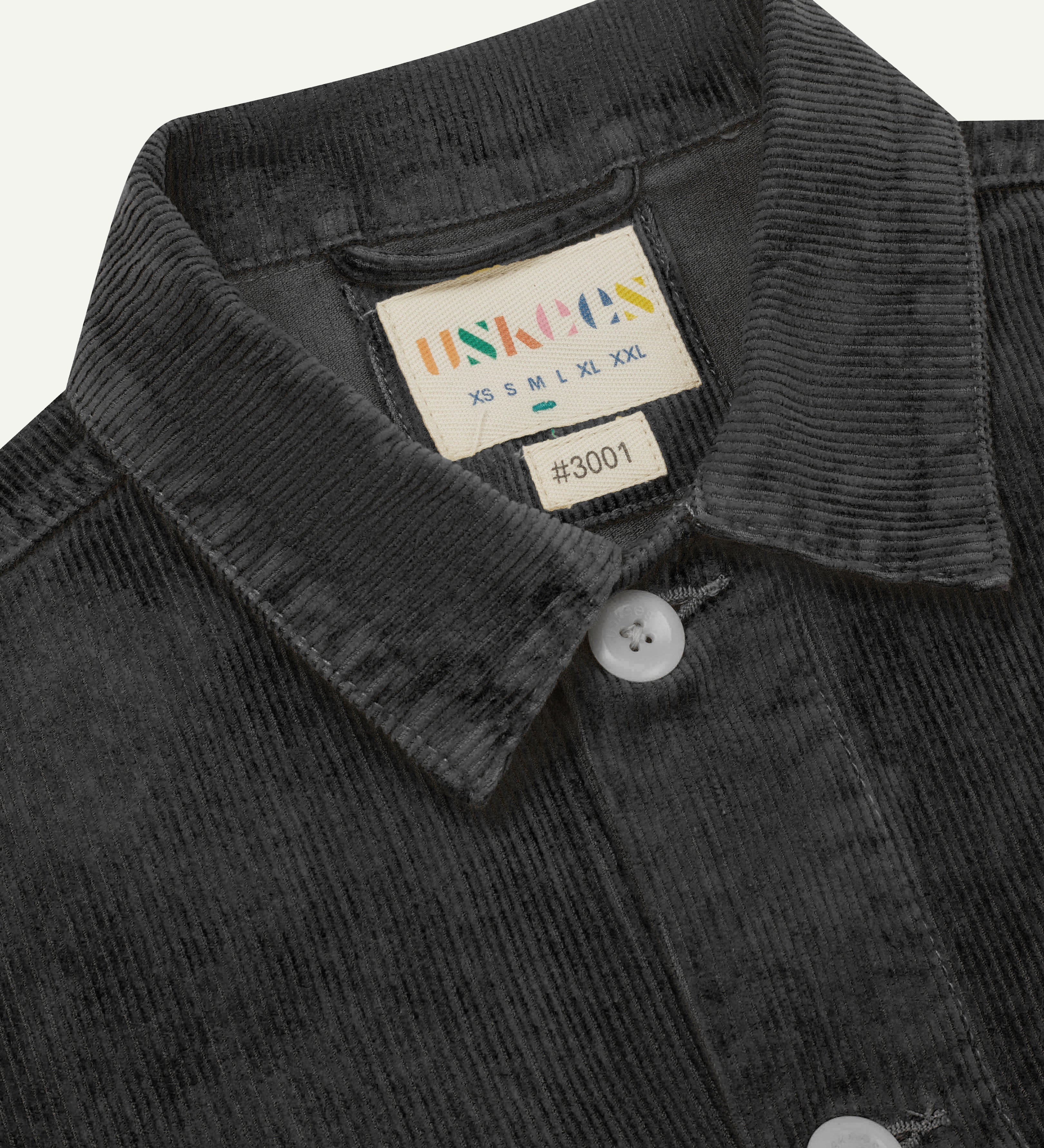 Close-up view of dark grey, buttoned corduroy overshirt from Uskees. Clear view of corozo buttons, uskees brand label and collar.