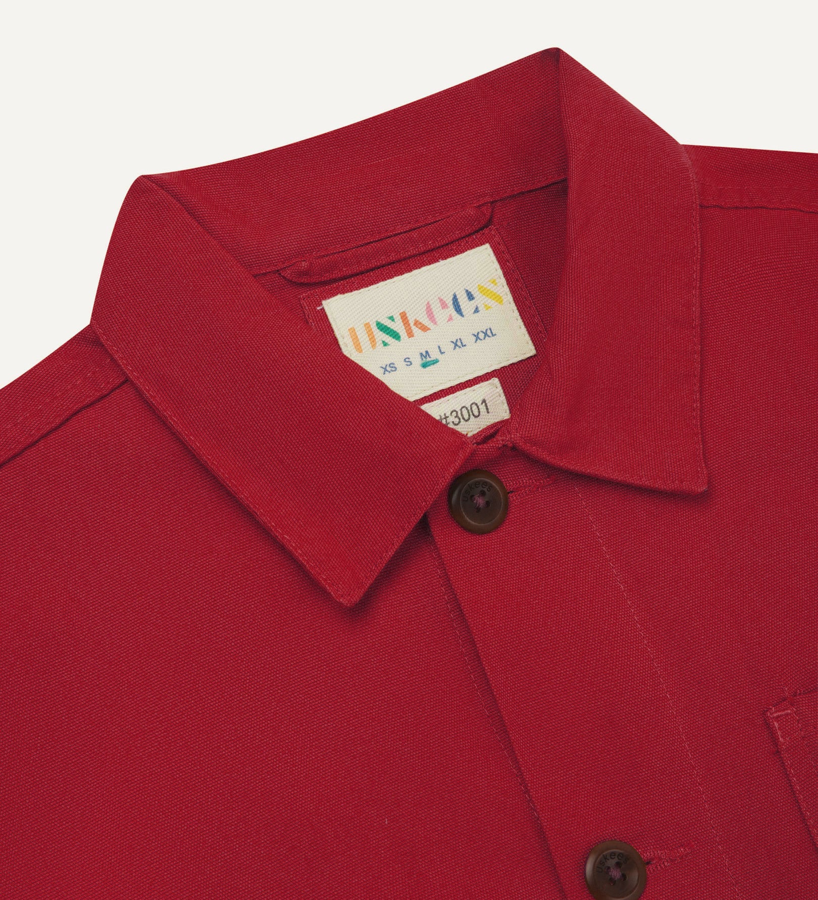 Close-up view of 3001 berry red, buttoned organic cotton overshirt from Uskees showing corozo buttons, branding label, collar and hanging loop.