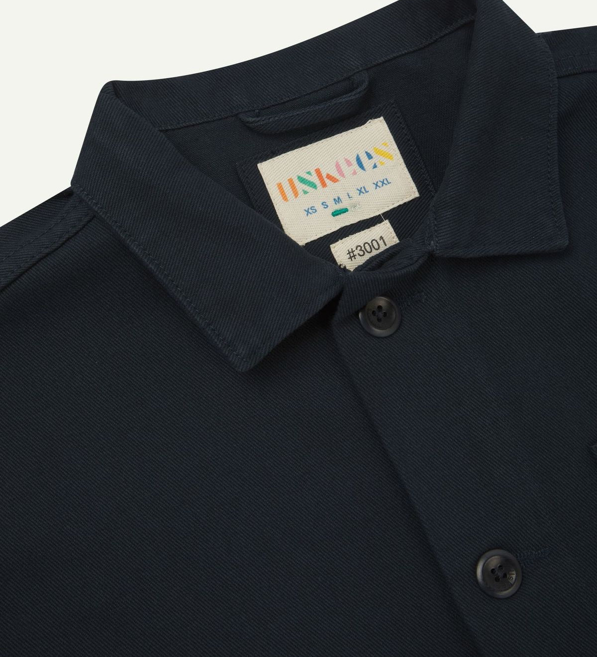 Close-up top-half view of #3001, dark blue (blueberry) organic cotton drill overshirt. With focus on collar, Uskees brand label and corozo buttons.