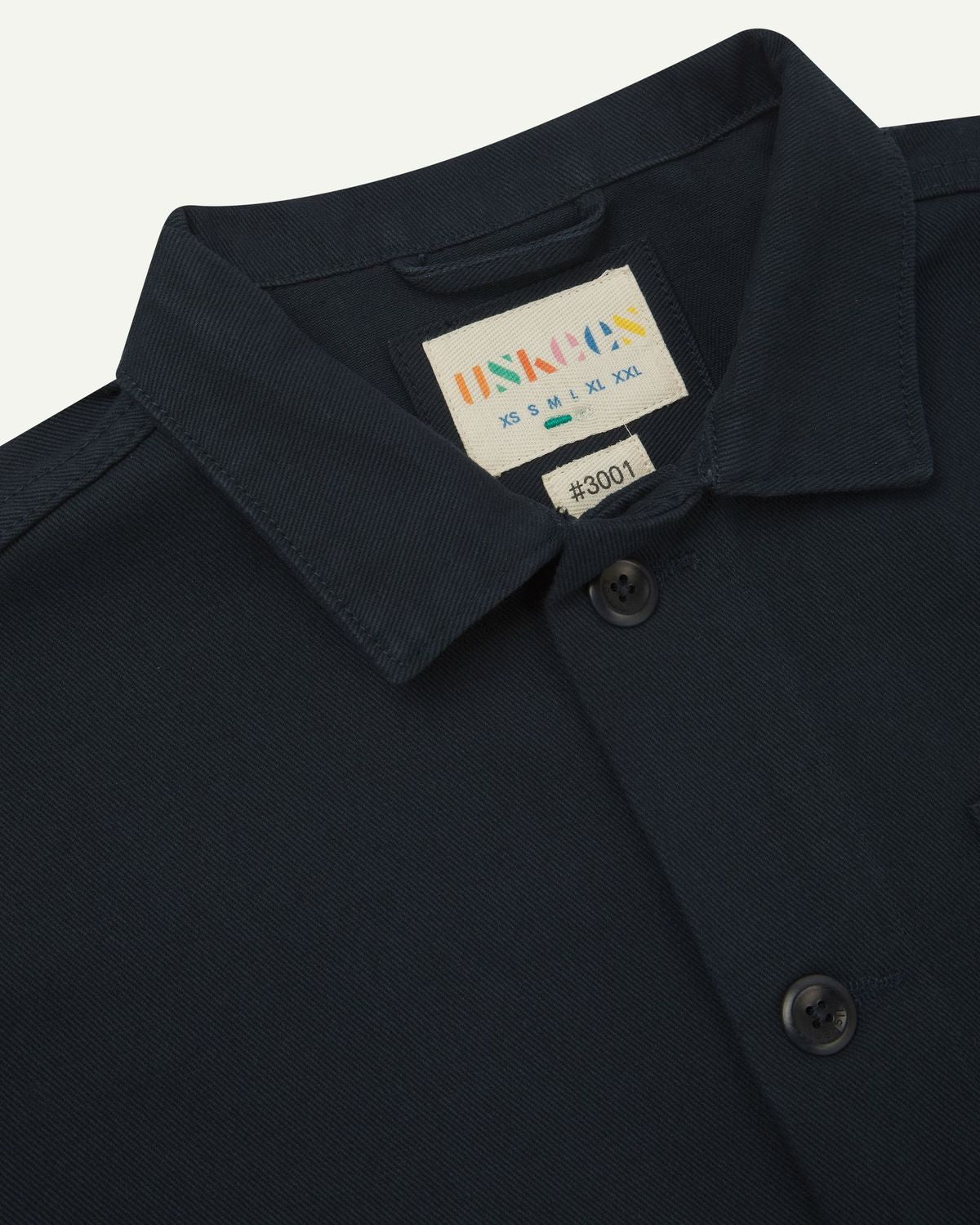 Close-up top-half view of #3001, dark blue (blueberry) organic cotton drill overshirt. With focus on collar, Uskees brand label and corozo buttons.