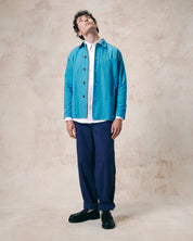 Male model wearing bright blue, unbuttoned organic cotton overshirt. Clear view of chest and hip pockets, corozo buttons and Uskees trousers with cuffs slightly rolled up.