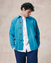 Front model shot of bright blue, unbuttoned organic cotton overshirt over a white buttoned up shirt. Clear view of overshirt chest and hip pockets and corozo buttons