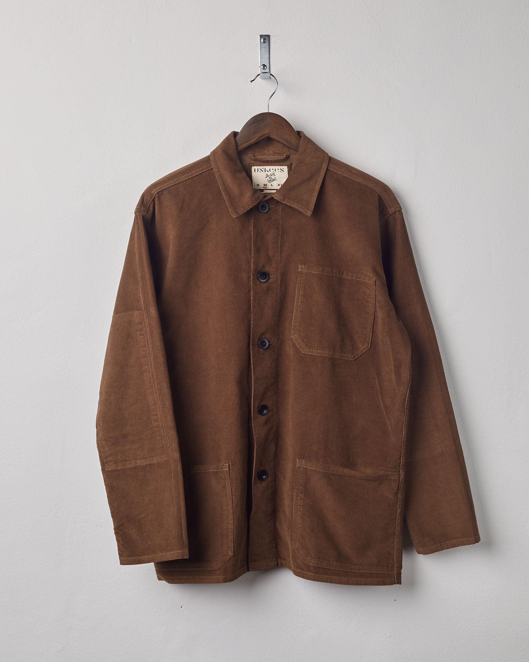 Brown coloured, buttoned corduroy overshirt from Uskees presented on hanger. Clear view of breast and hip pocket and corozo buttons.