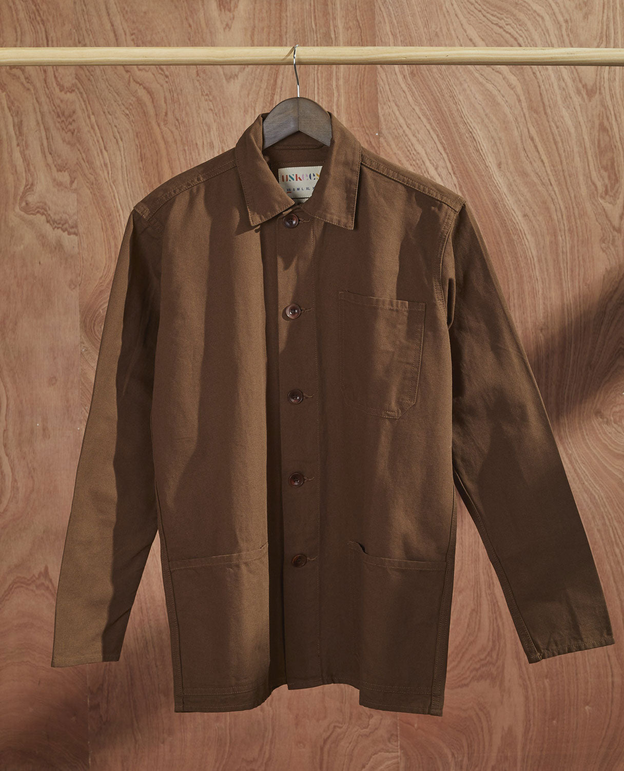 Front-view of brown buttoned overshirt from Uskees on hanger. Clear view of breast and hip pocket and corozo buttons.