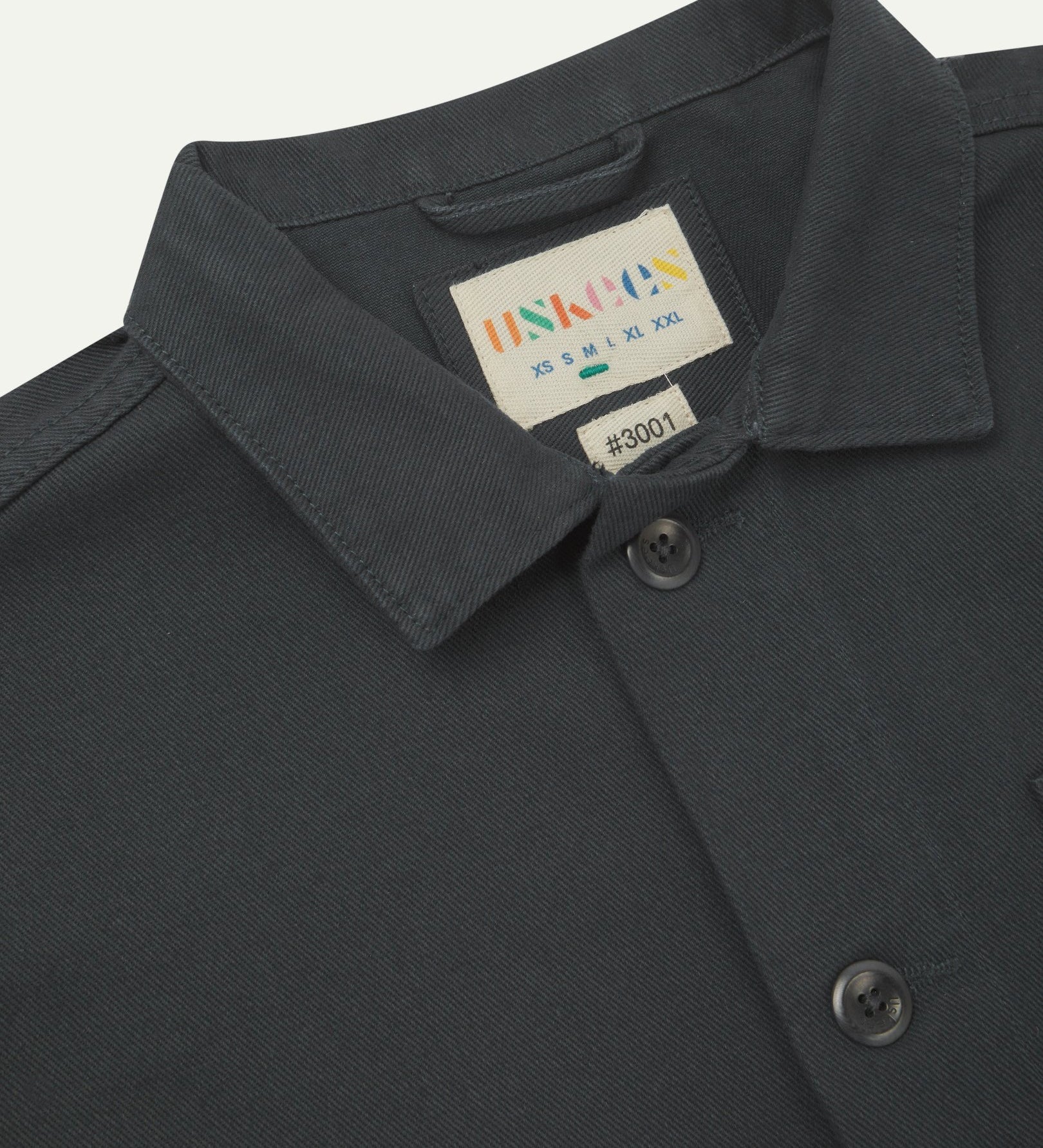 Close-up top-half view of #3001, charcoal-grey organic cotton drill overshirt. With focus on collar, Uskees brand label and corozo buttons.