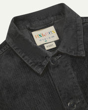 Close-up view of 3001 charcoal-grey, buttoned corduroy overshirt from Uskees showing corozo buttons, brand/size label, collar and hanging hoop.