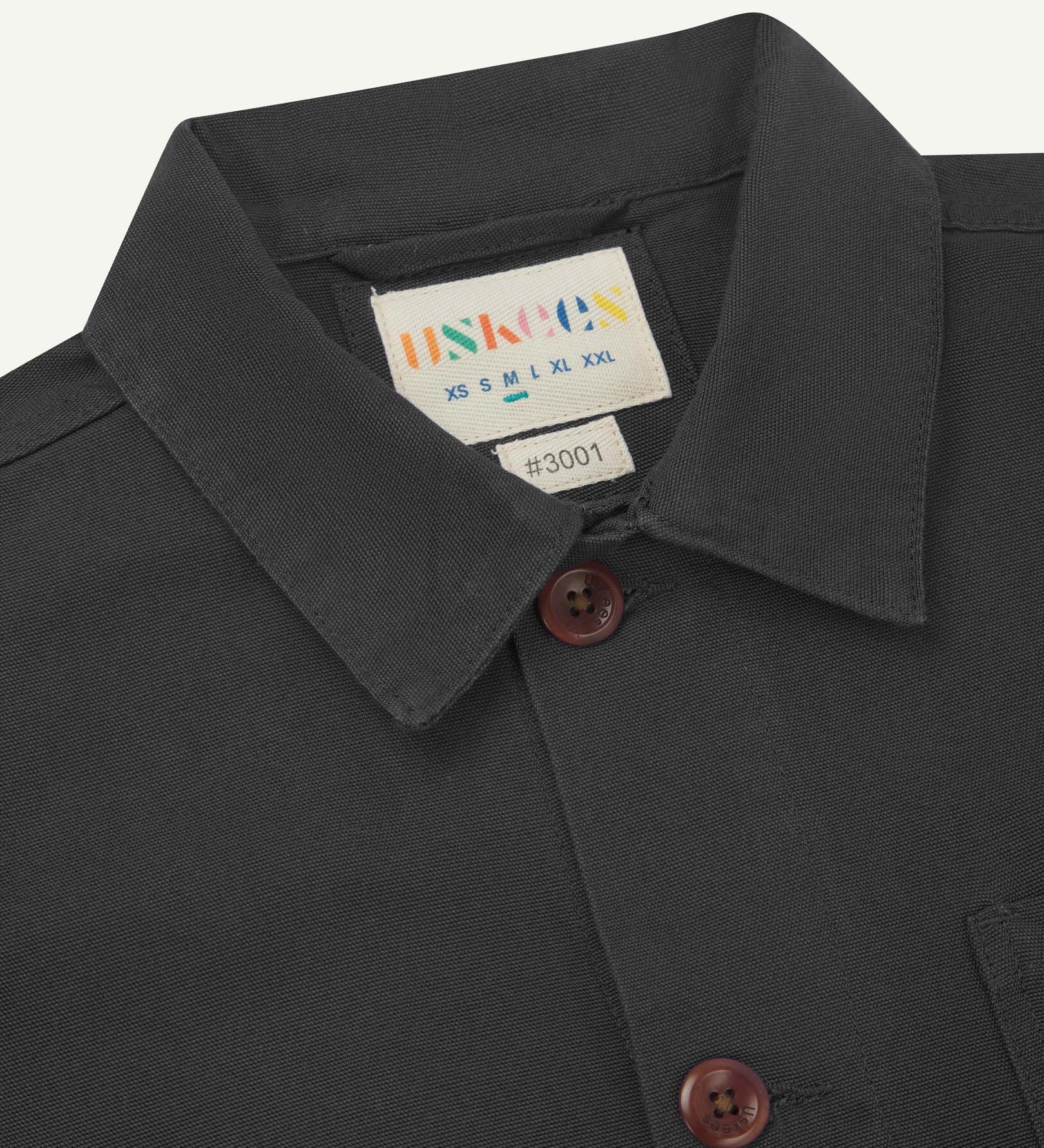 Close-up view of 3001 charcoal-grey, buttoned organic cotton overshirt from Uskees showing corozo buttons, brand label, collar and hanging hoop.