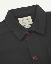 Close-up view of 3001 charcoal-grey, buttoned organic cotton overshirt from Uskees showing corozo buttons, brand label, collar and hanging hoop.