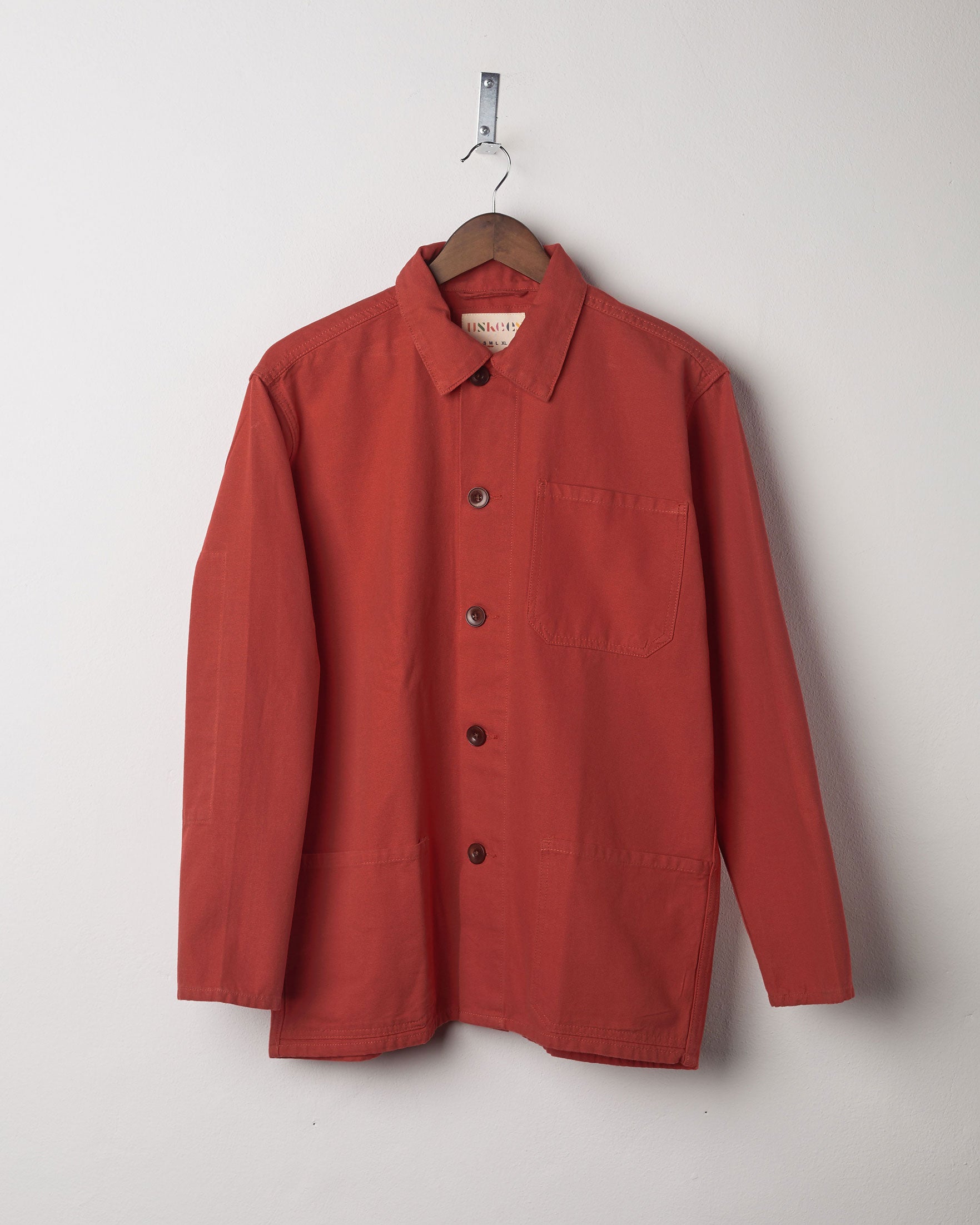 Clay coloured, buttoned overshirt from Uskees presented on hanger. Clear view of breast and hip pocket and corozo buttons.