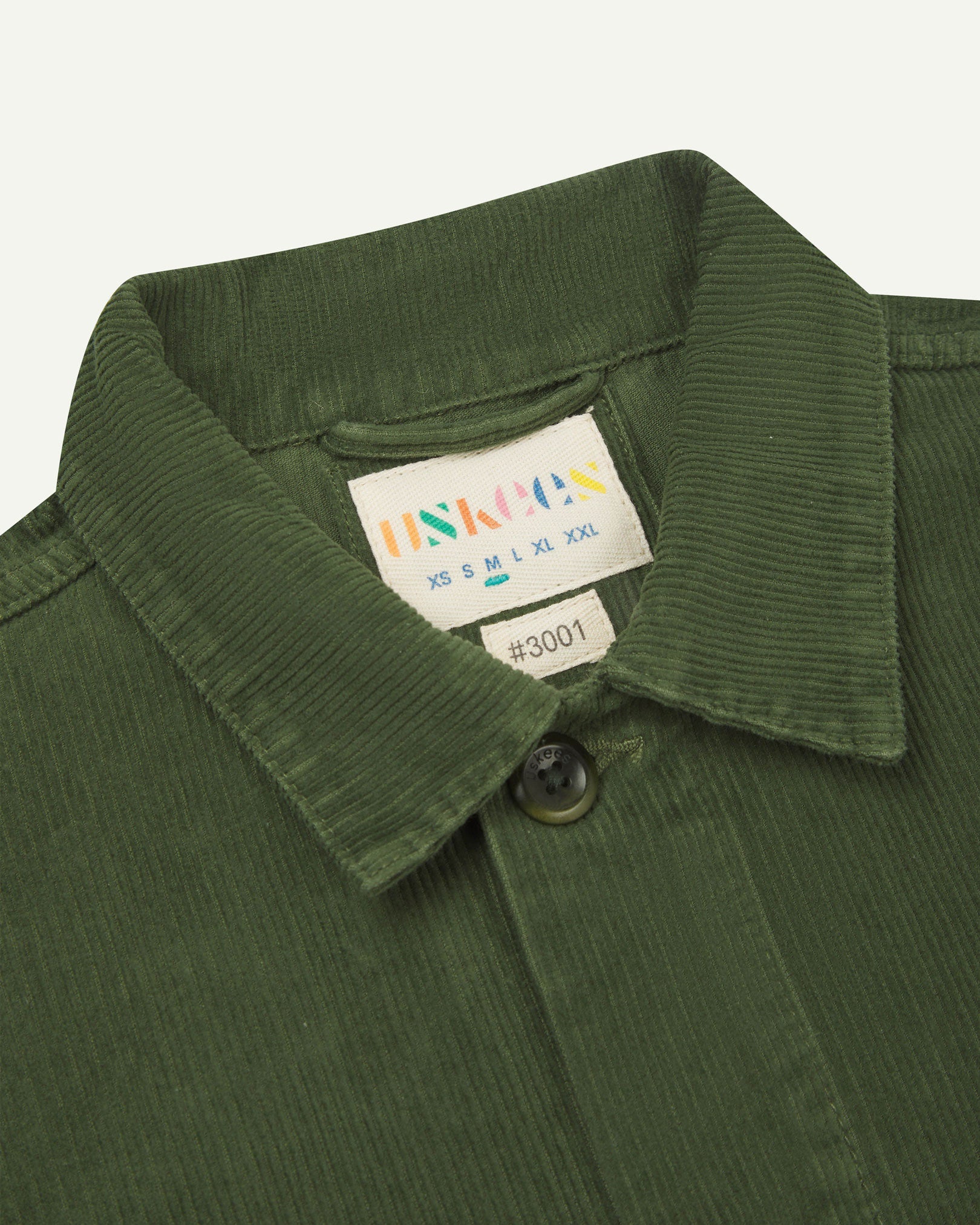 Close-up view of 3001 coriander-green, buttoned corduroy overshirt from Uskees showing corozo buttons, brand/size label, collar and hanging hoop.