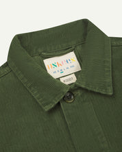 Close-up view of 3001 coriander-green, buttoned corduroy overshirt from Uskees showing corozo buttons, brand/size label, collar and hanging hoop.