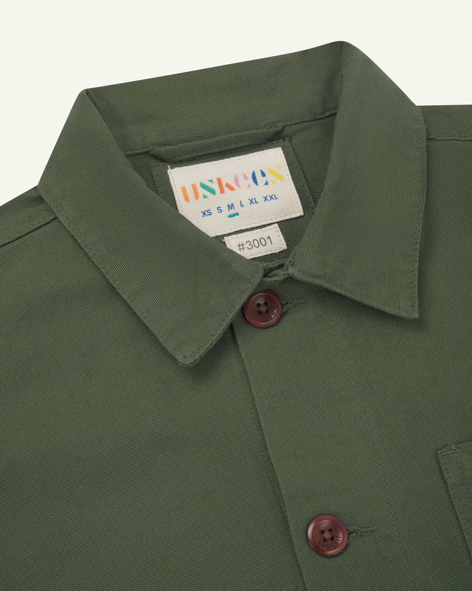 Close-up view of 3001 coriander-green, buttoned organic cotton overshirt from Uskees showing corozo buttons, brand label, collar and hanging hoop.