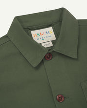 Close-up view of 3001 coriander-green, buttoned organic cotton overshirt from Uskees showing corozo buttons, brand label, collar and hanging hoop.