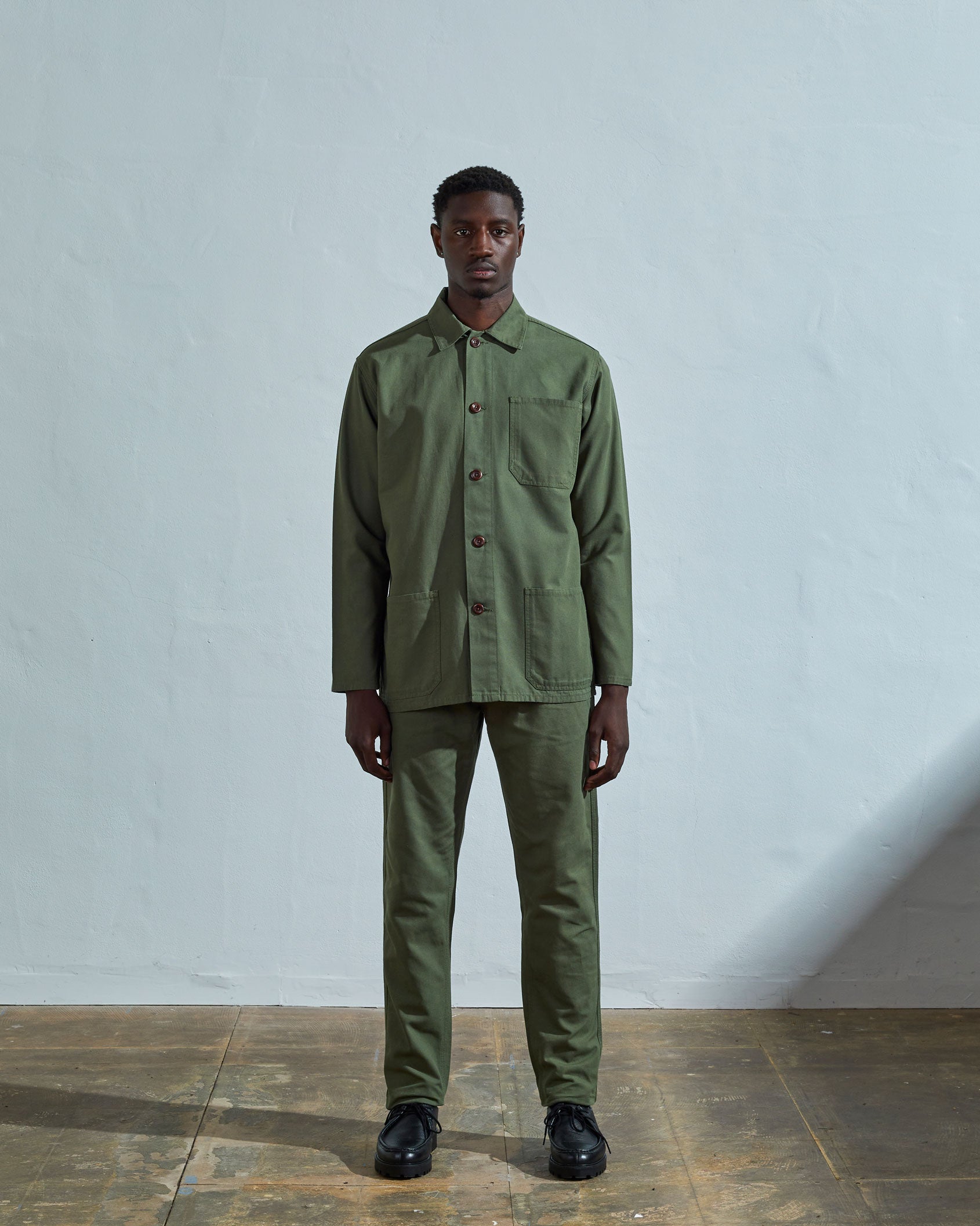 Full-length front view of model wearing #3001, coriander-green organic cotton overshirt paired with dark pants.