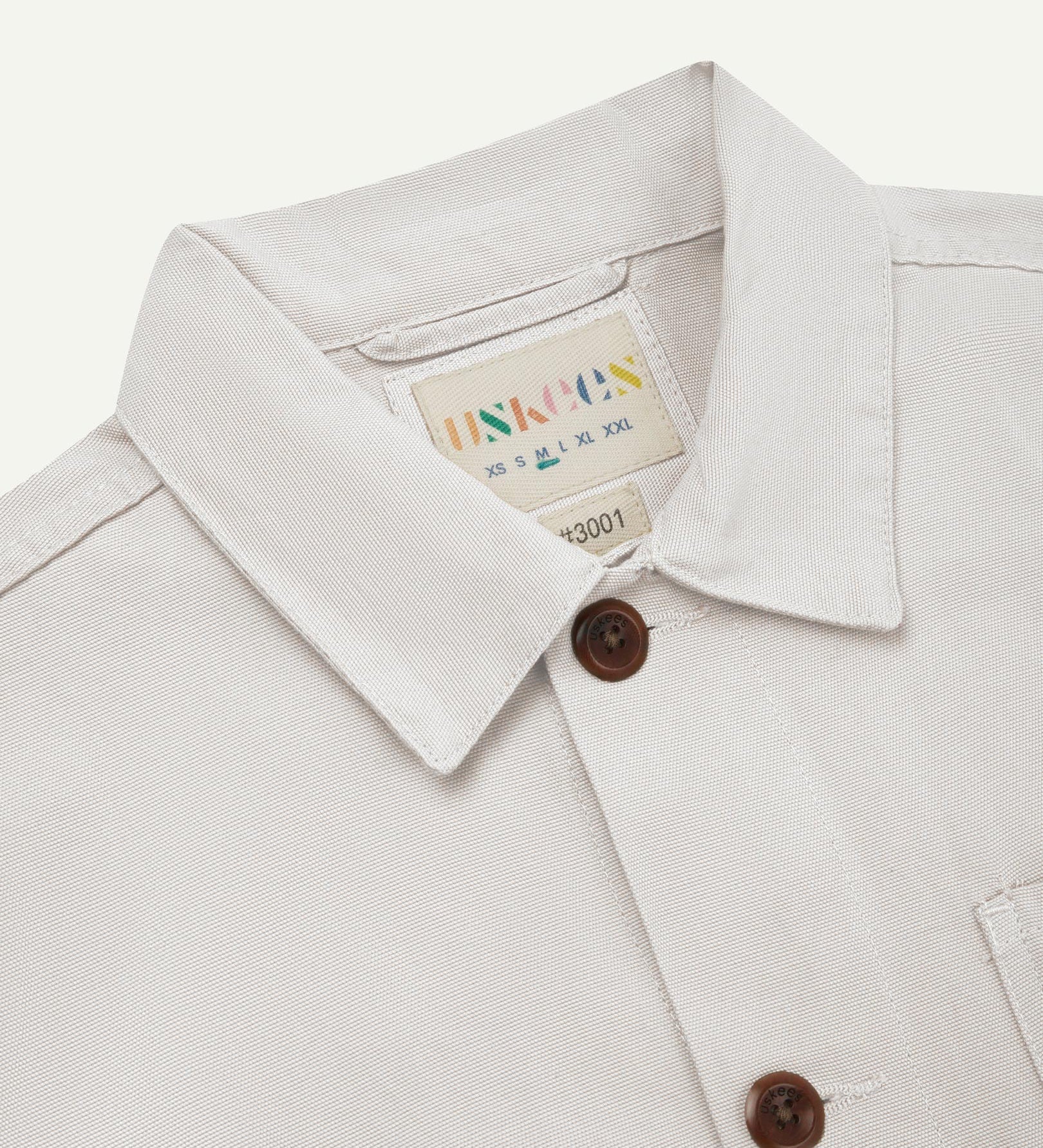 Close-up view of 3001 cream, buttoned organic cotton overshirt from Uskees showing corozo buttons, brand label, collar and hanging hoop.