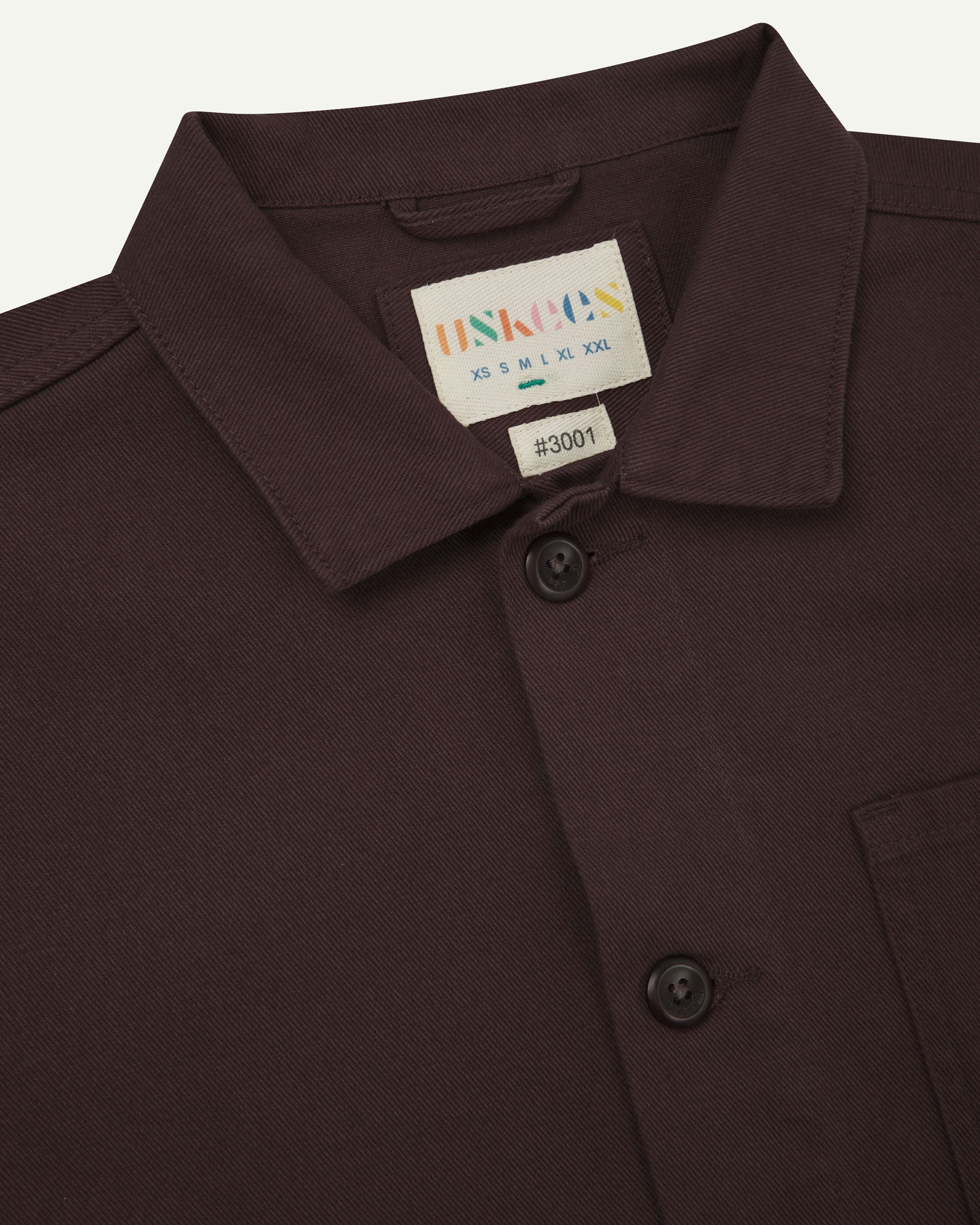 Close-up top-half view of #3001, burgundy-brown organic cotton drill overshirt. With focus on collar, Uskees brand label and corozo buttons.