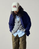 Male model wearing a blue overshirt, gray cardigan, light blue shirt, and brown pants with a cream cap featuring 'USK' on a plain background.