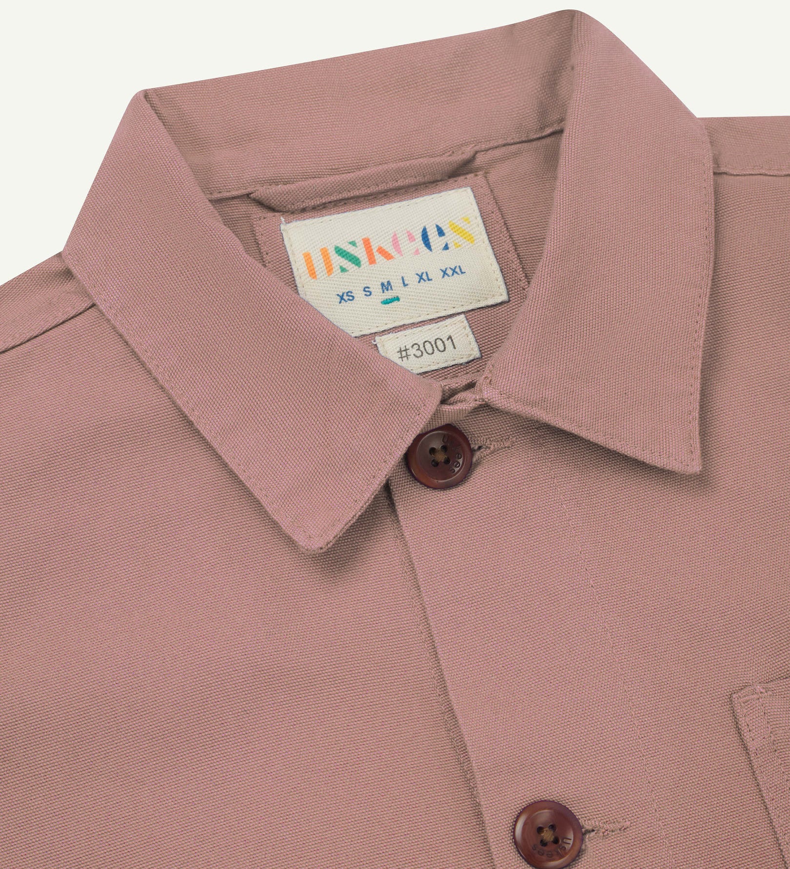 Close-up view of 3001 dusty pink, buttoned organic cotton overshirt from Uskees showing corozo buttons, brand label, collar and hanging hoop.
