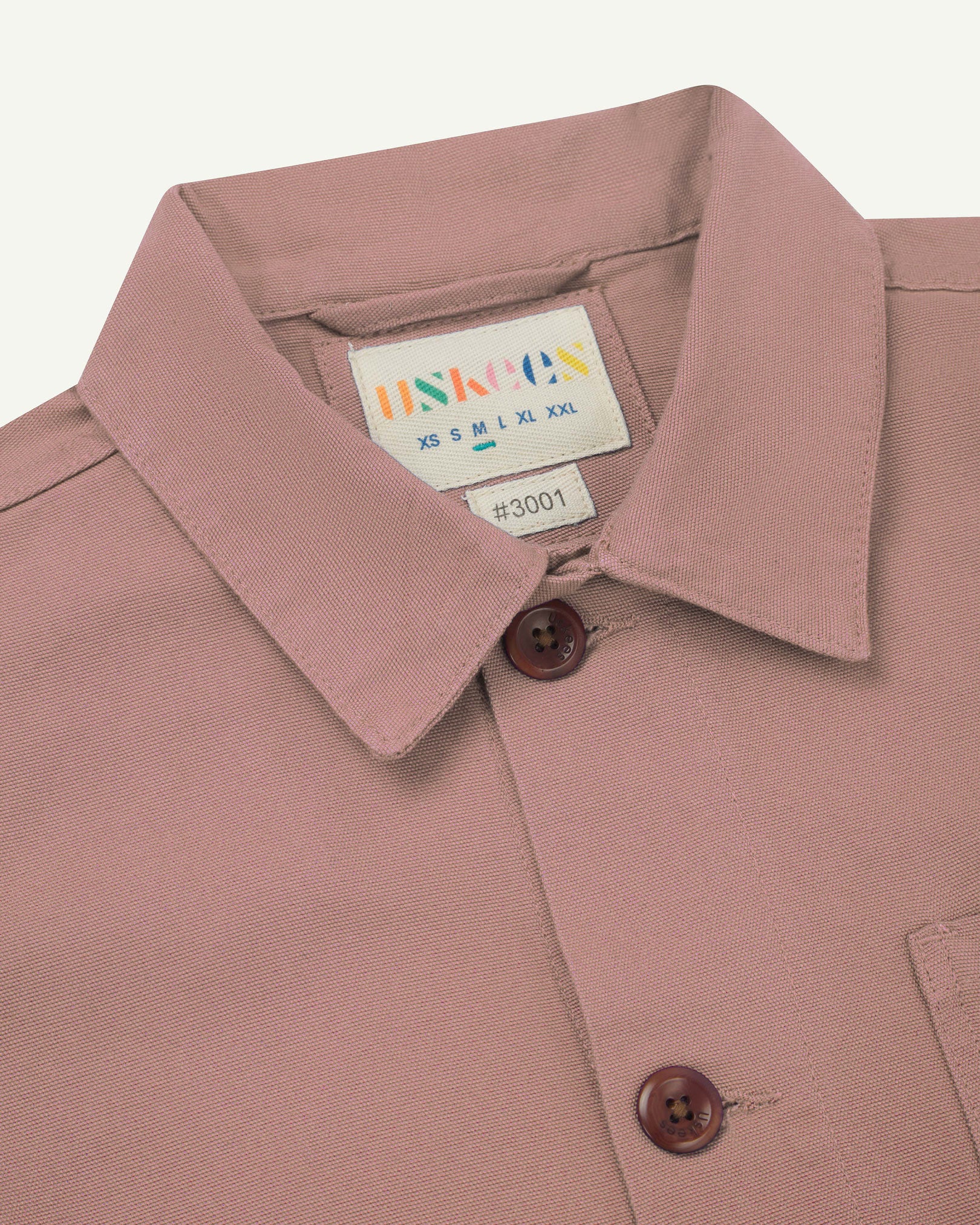 Close-up view of 3001 dusty pink, buttoned organic cotton overshirt from Uskees showing corozo buttons, brand label, collar and hanging hoop.