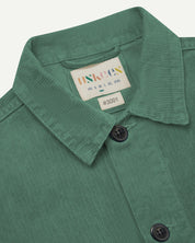 Close-up view of 3001 light green, buttoned corduroy overshirt from Uskees showing corozo buttons, brand/size label, collar and hanging hoop.