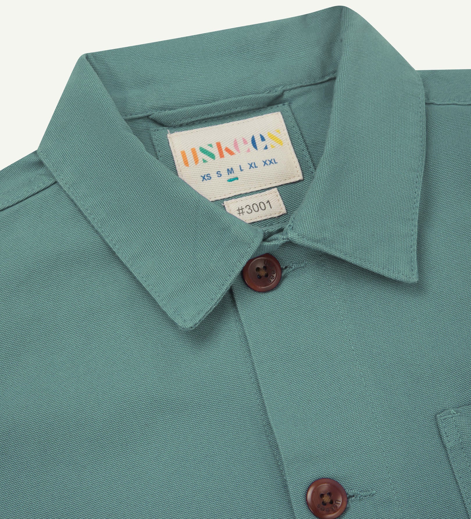 Close-up view of 3001 blue-green (eucalyptus), buttoned organic cotton overshirt from Uskees showing corozo buttons, brand label, collar and hanging hoop.