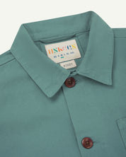 Close-up view of 3001 blue-green (eucalyptus), buttoned organic cotton overshirt from Uskees showing corozo buttons, brand label, collar and hanging hoop.