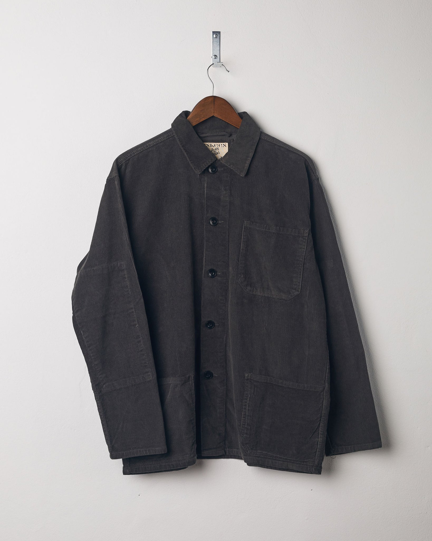 Faded black coloured, buttoned corduroy overshirt from Uskees presented on hanger. Clear view of breast and hip pocket and corozo buttons.
