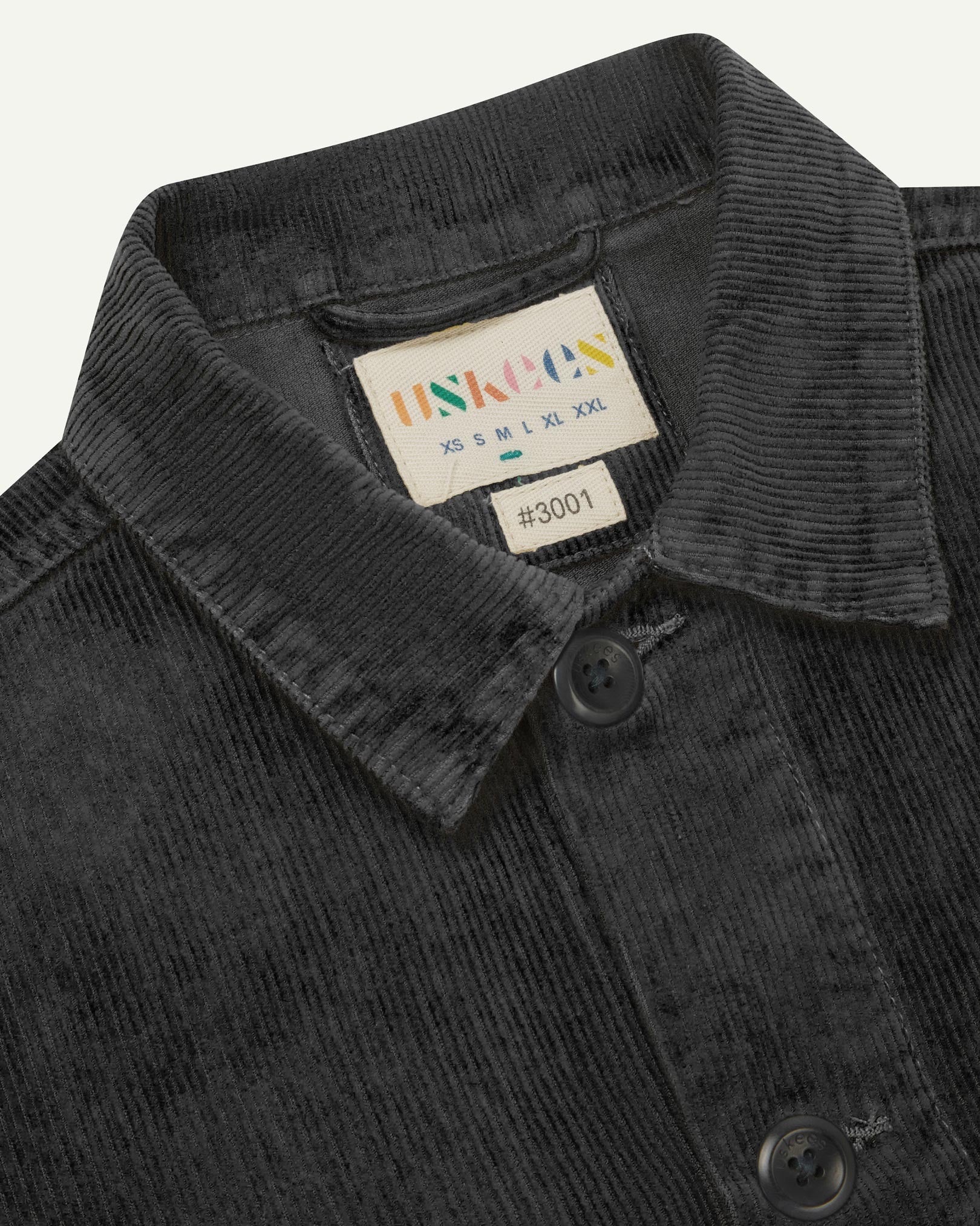 Close-up view of 3001 faded black, buttoned corduroy overshirt from Uskees showing corozo buttons, brand/size label, collar and hanging hoop.