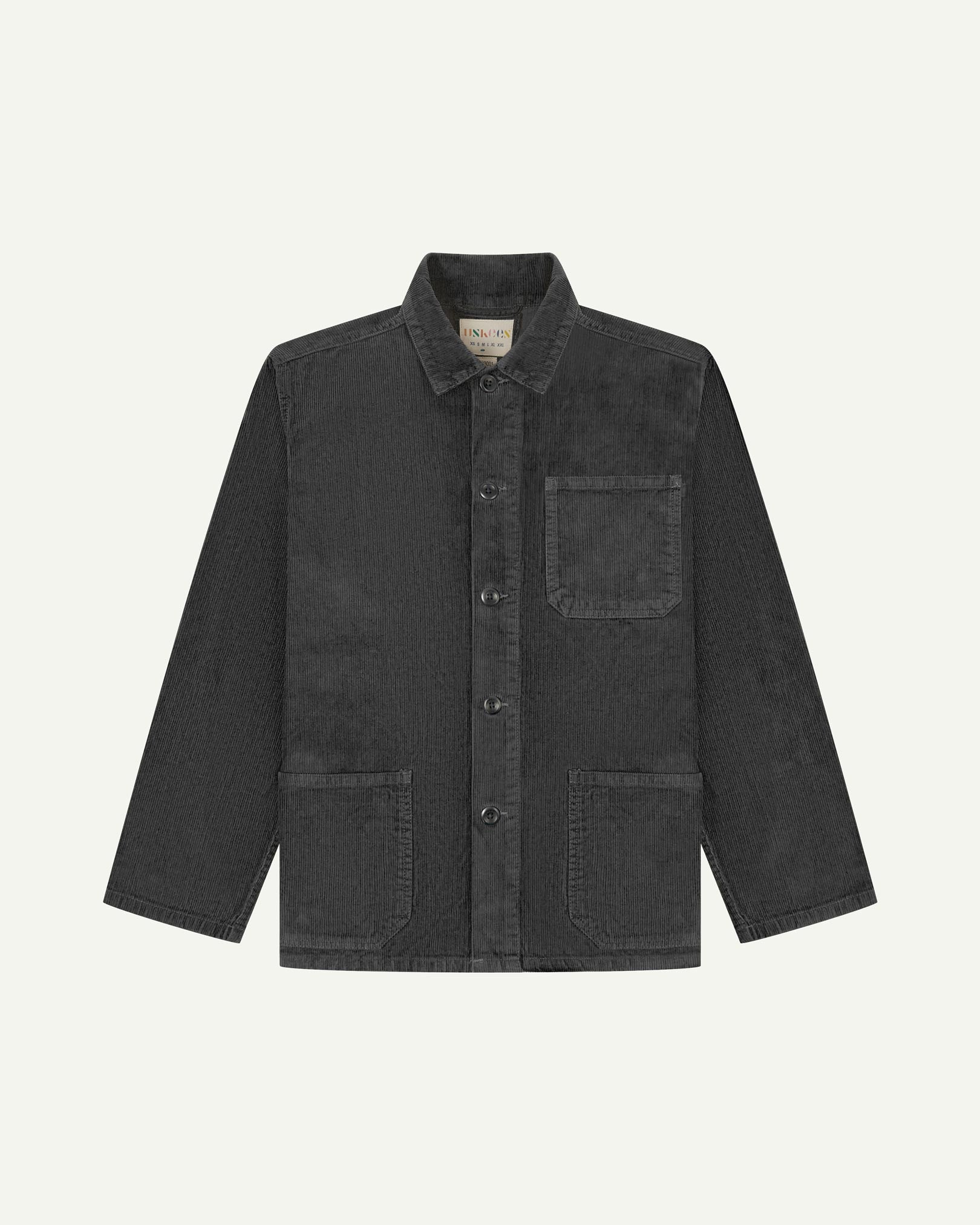 Front flat shot of faded black, buttoned corduroy overshirt. Clear view of chest and hip pockets, corozo buttons and Uskees branding label.