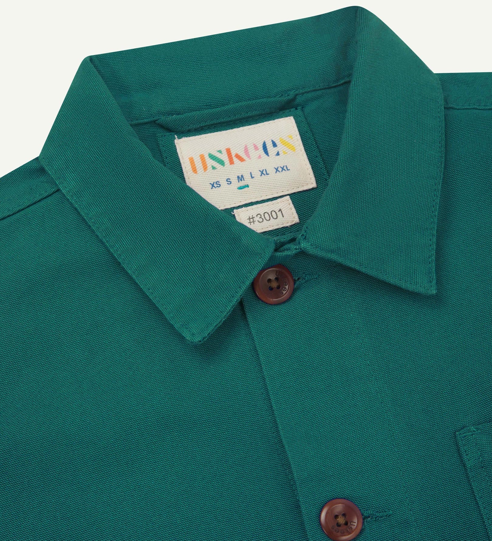 Close-up view of 3001 foam green, buttoned organic cotton overshirt from Uskees showing corozo buttons, brand label, collar and hanging hoop.