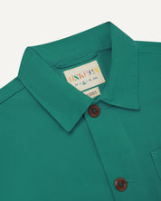 Close-up view of 3001 foam green, buttoned organic cotton overshirt from Uskees showing corozo buttons, brand label, collar and hanging hoop.