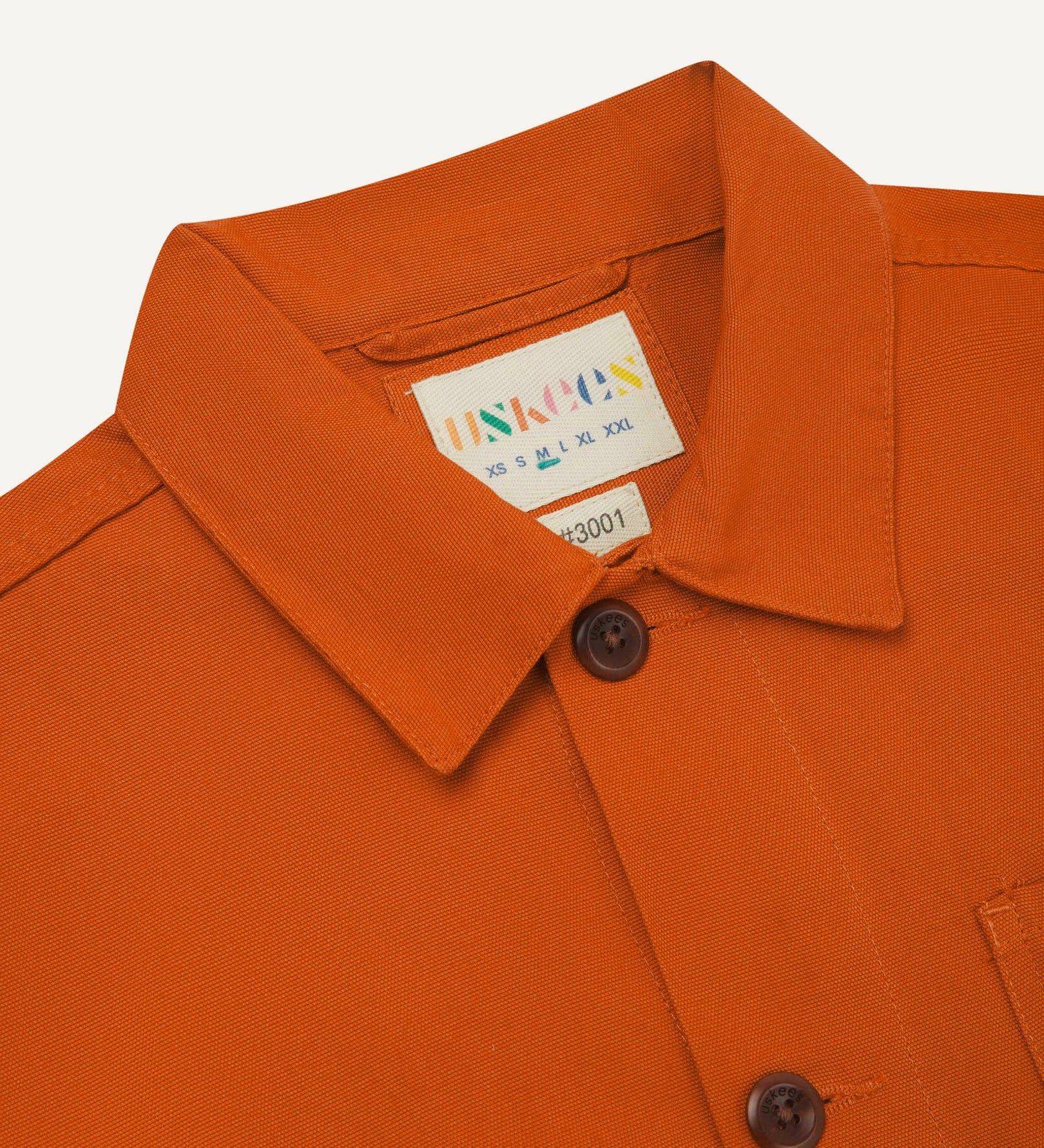 Close-up view of 3001 golden-orange, buttoned organic cotton overshirt from Uskees showing corozo buttons, brand label, collar and hanging hoop.