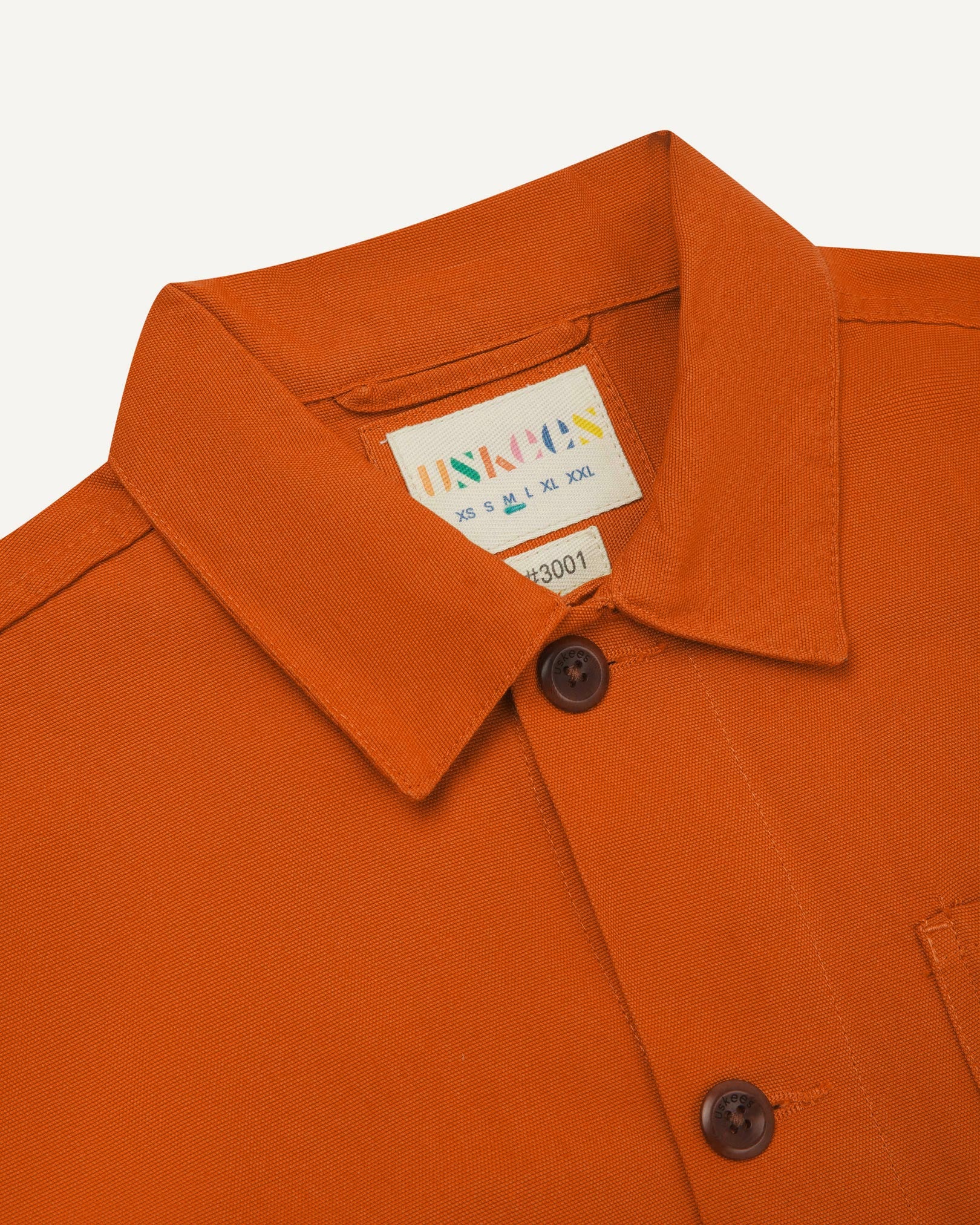 Close-up view of 3001 golden-orange, buttoned organic cotton overshirt from Uskees showing corozo buttons, brand label, collar and hanging hoop.