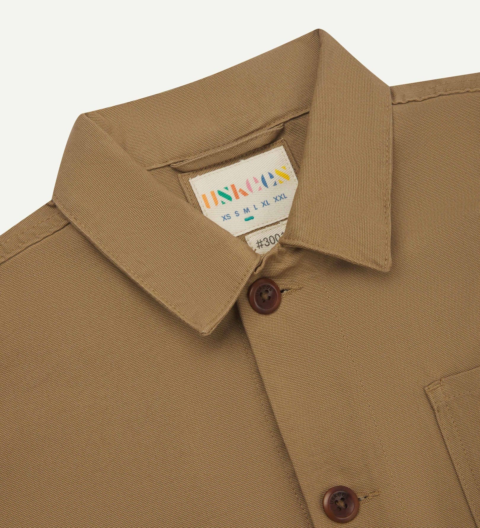 Close-up view of 3001 khaki, buttoned organic cotton overshirt from Uskees showing corozo buttons, brand label, collar and hanging hoop.