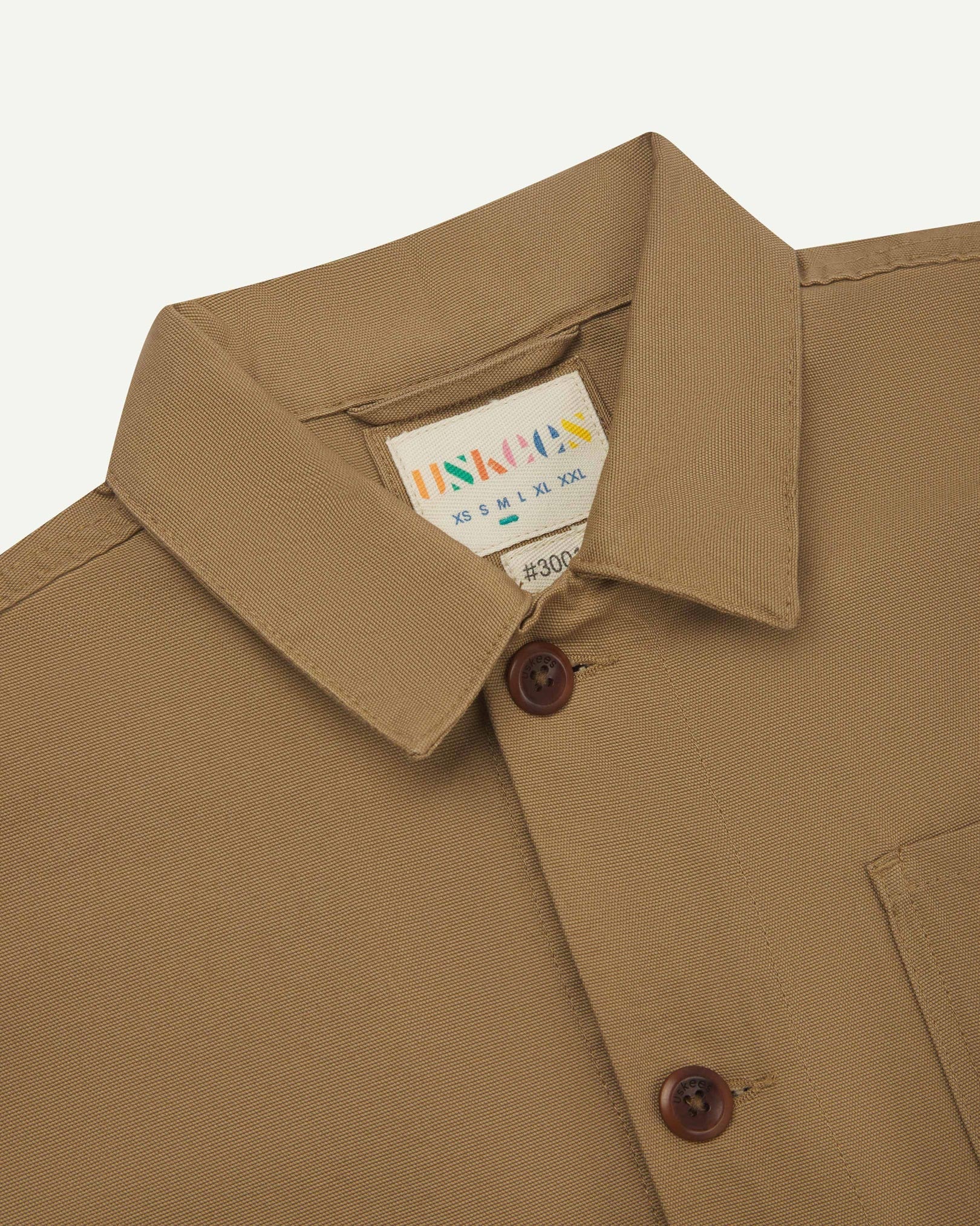 Close-up view of 3001 khaki, buttoned organic cotton overshirt from Uskees showing corozo buttons, brand label, collar and hanging hoop.