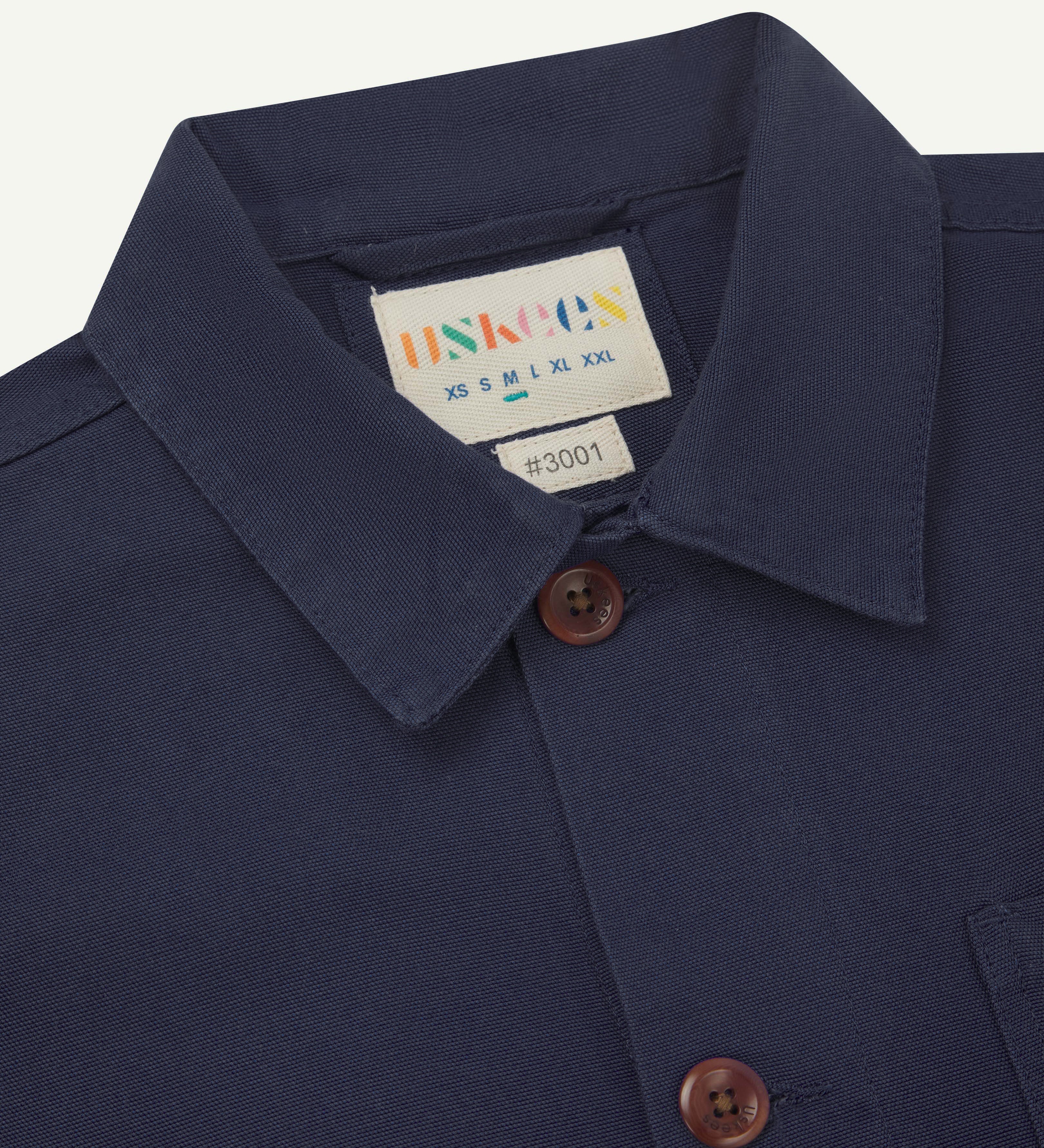 Close-up view of 3001 midnight blue, buttoned organic cotton overshirt from Uskees showing corozo buttons, brand label, collar and hanging hoop.