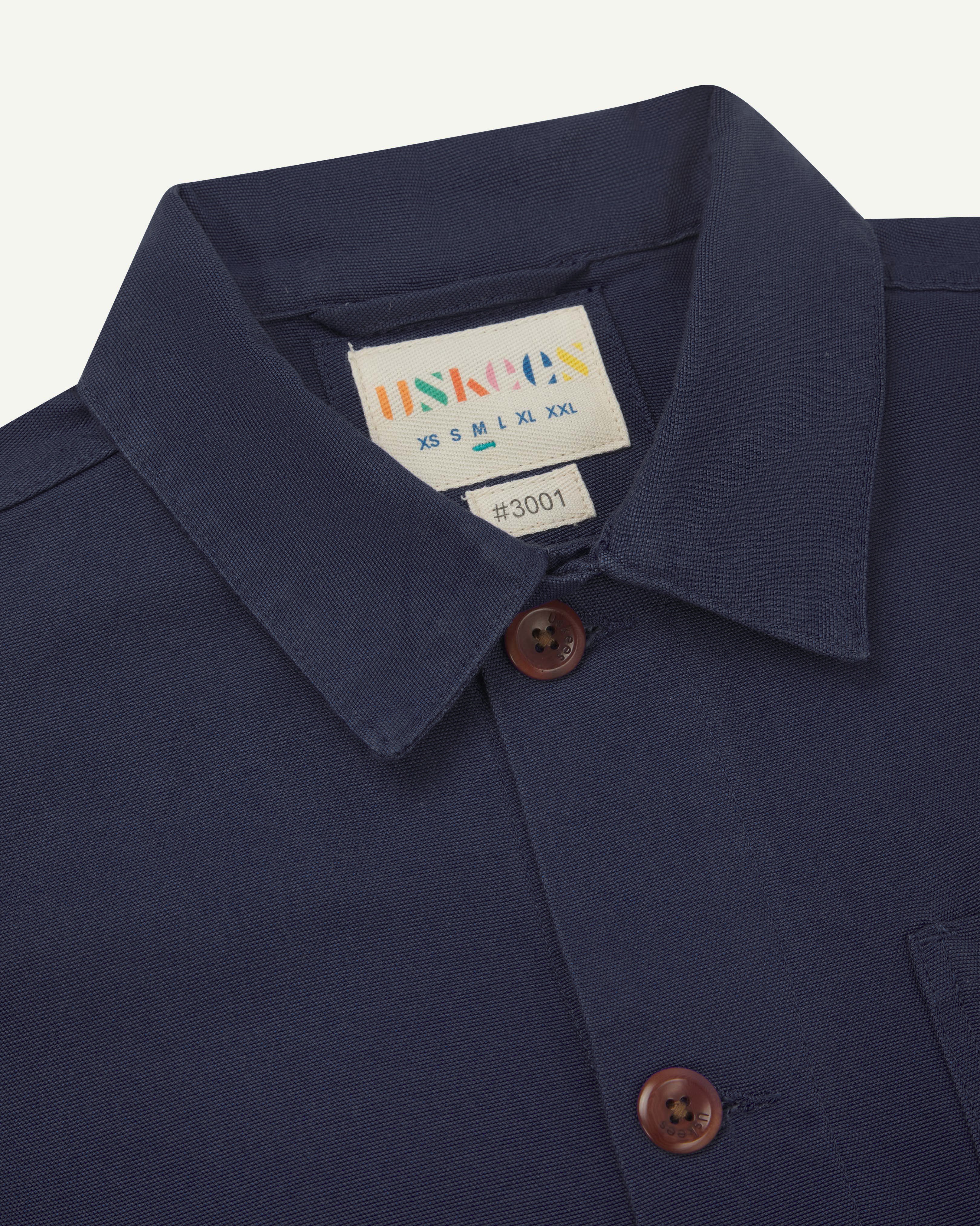 Close-up view of 3001 midnight blue, buttoned organic cotton overshirt from Uskees showing corozo buttons, brand label, collar and hanging hoop.