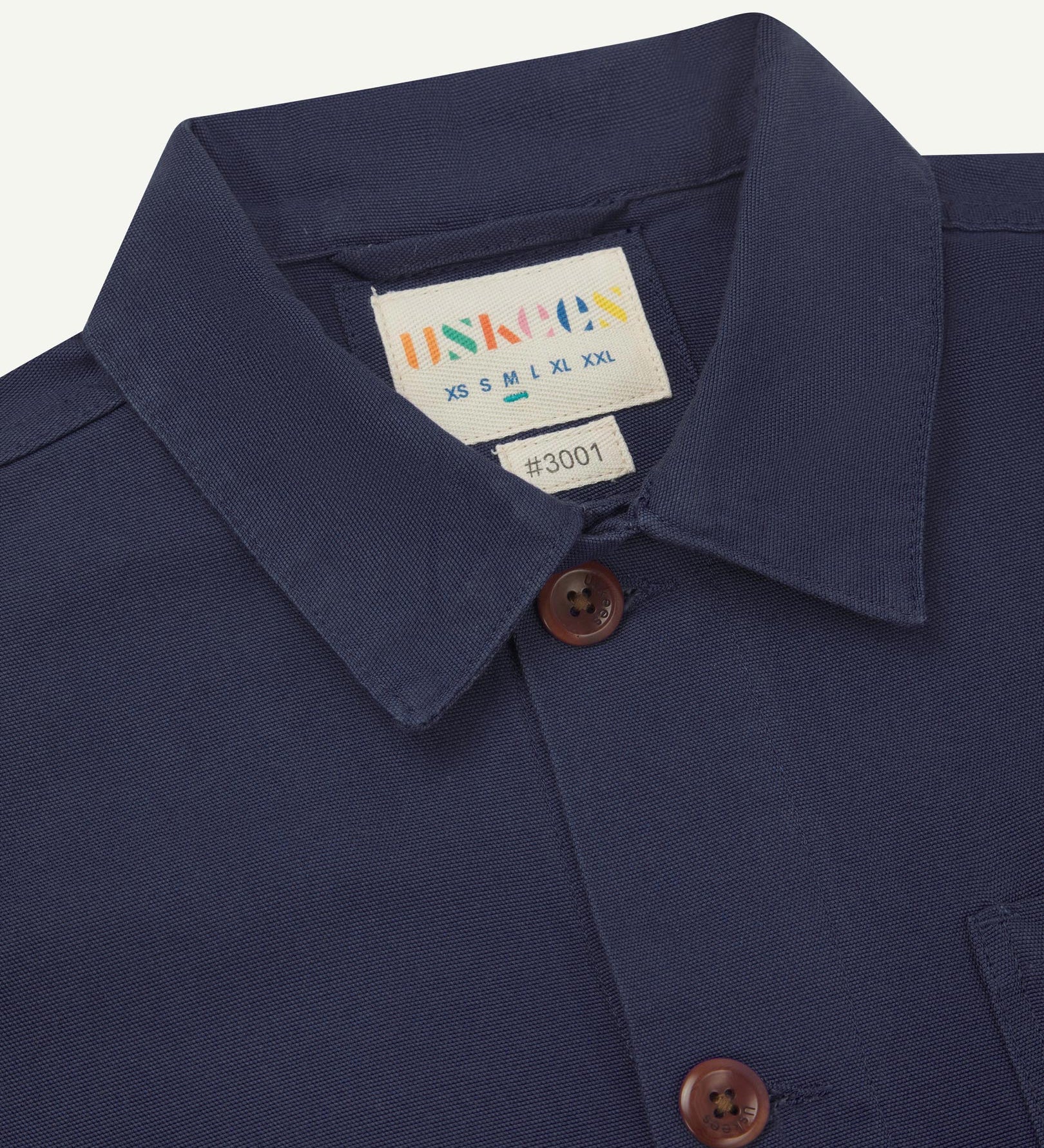 Close-up view of 3001 midnight blue, buttoned organic cotton overshirt from Uskees showing corozo buttons, brand label, collar and hanging hoop.